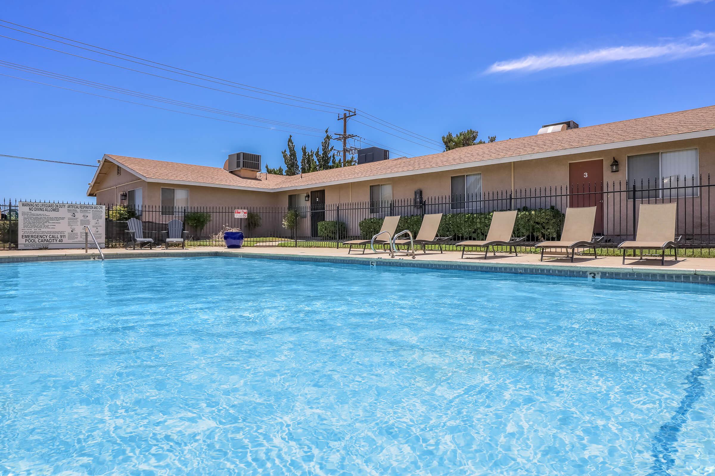 A clear blue swimming pool in front of a simple one-story apartment building. The pool is surrounded by lounge chairs and has a fence in the background. Bright sunlight illuminates the scene, with a few trees visible and a clear blue sky overhead.