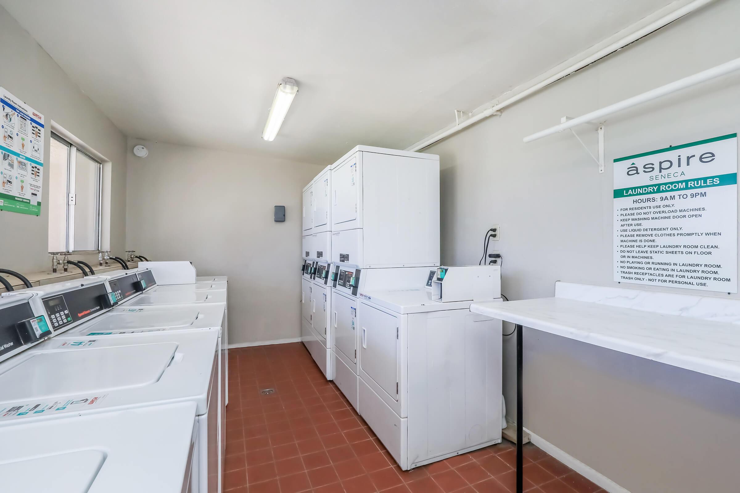 Interior of a laundry room featuring multiple white washing machines and dryers lined against the wall. A folding table is nearby, and a sign displaying laundry room rules is visible. The room is well-lit with fluorescent lights and has tiled flooring.