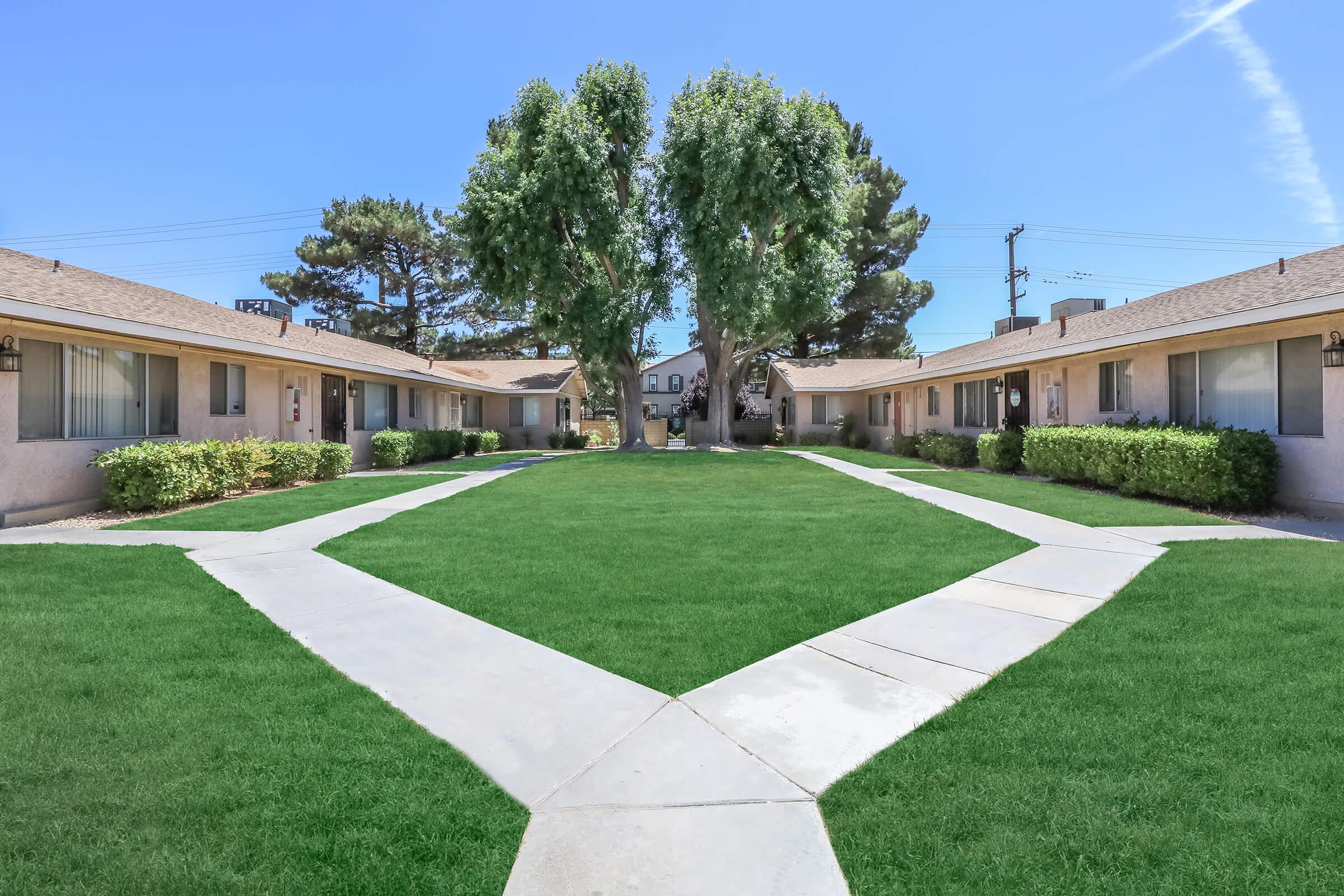 A view of a residential complex featuring a well-maintained grassy courtyard surrounded by two rows of single-story buildings. The pathway divides the lawn, leading to the entrance of the units. Tall trees provide shade, and the sky is clear and blue, creating a bright, inviting atmosphere.