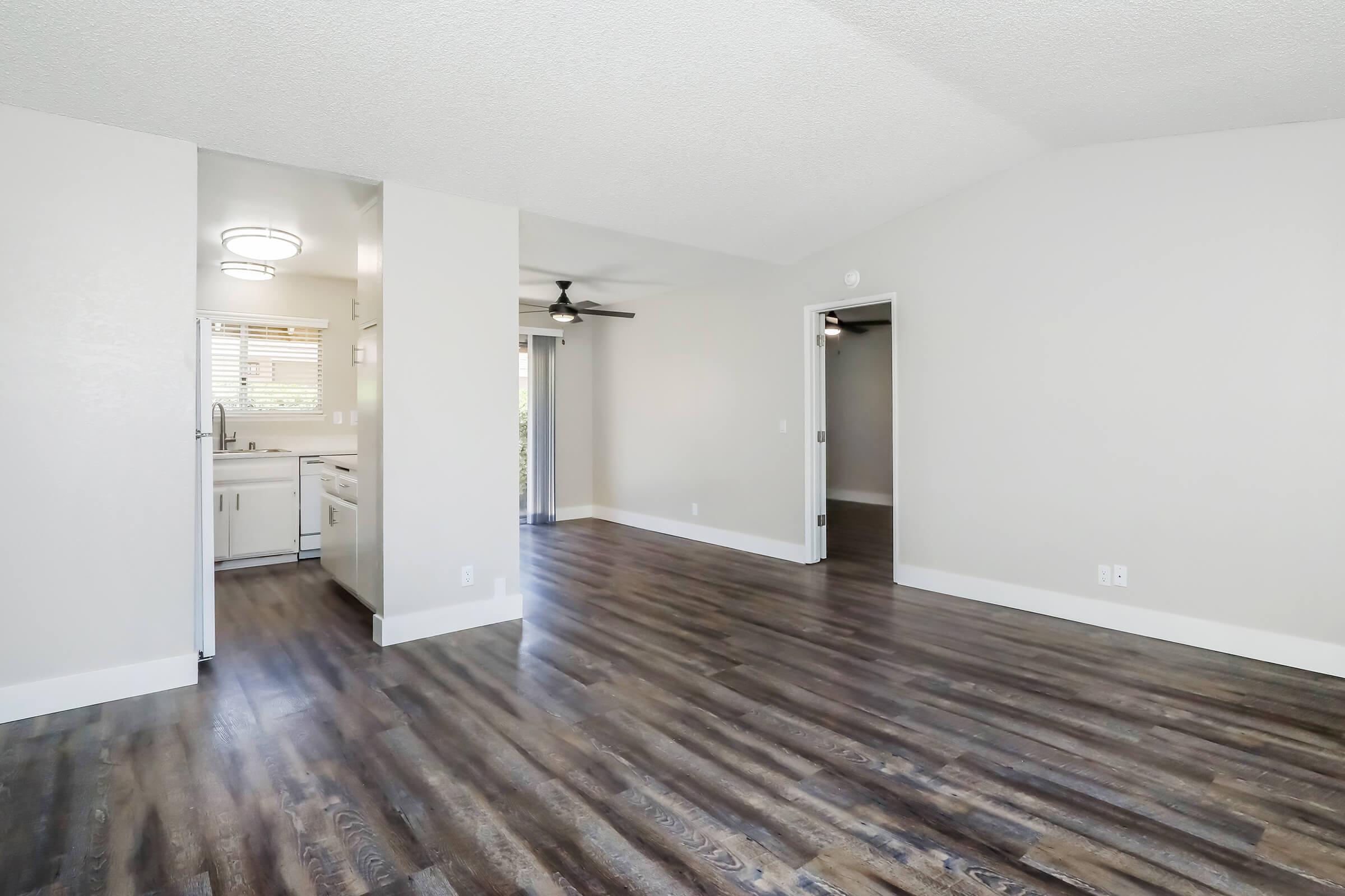 Spacious living area with hardwood-style flooring, features a ceiling fan and an open layout leading to the kitchen. Light-colored walls complement the natural light entering through the windows. A doorway on the right leads to another room, creating a cozy yet airy atmosphere.