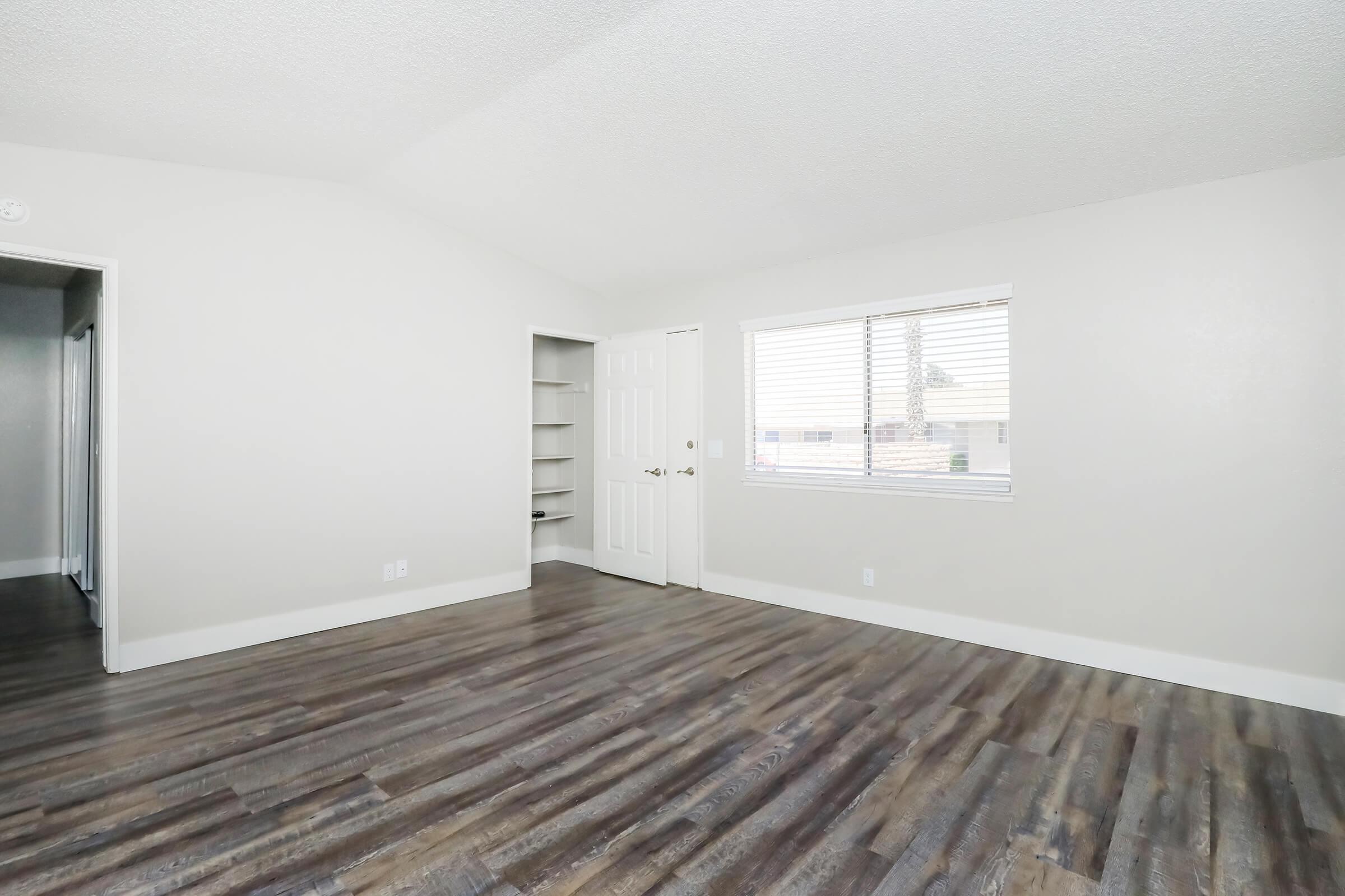 A well-lit room with gray walls and wood-like flooring. One wall features a window with blinds, allowing natural light in. A doorway leads to another room on the left, and a closet with shelves is visible in the back. The overall atmosphere is open and inviting.