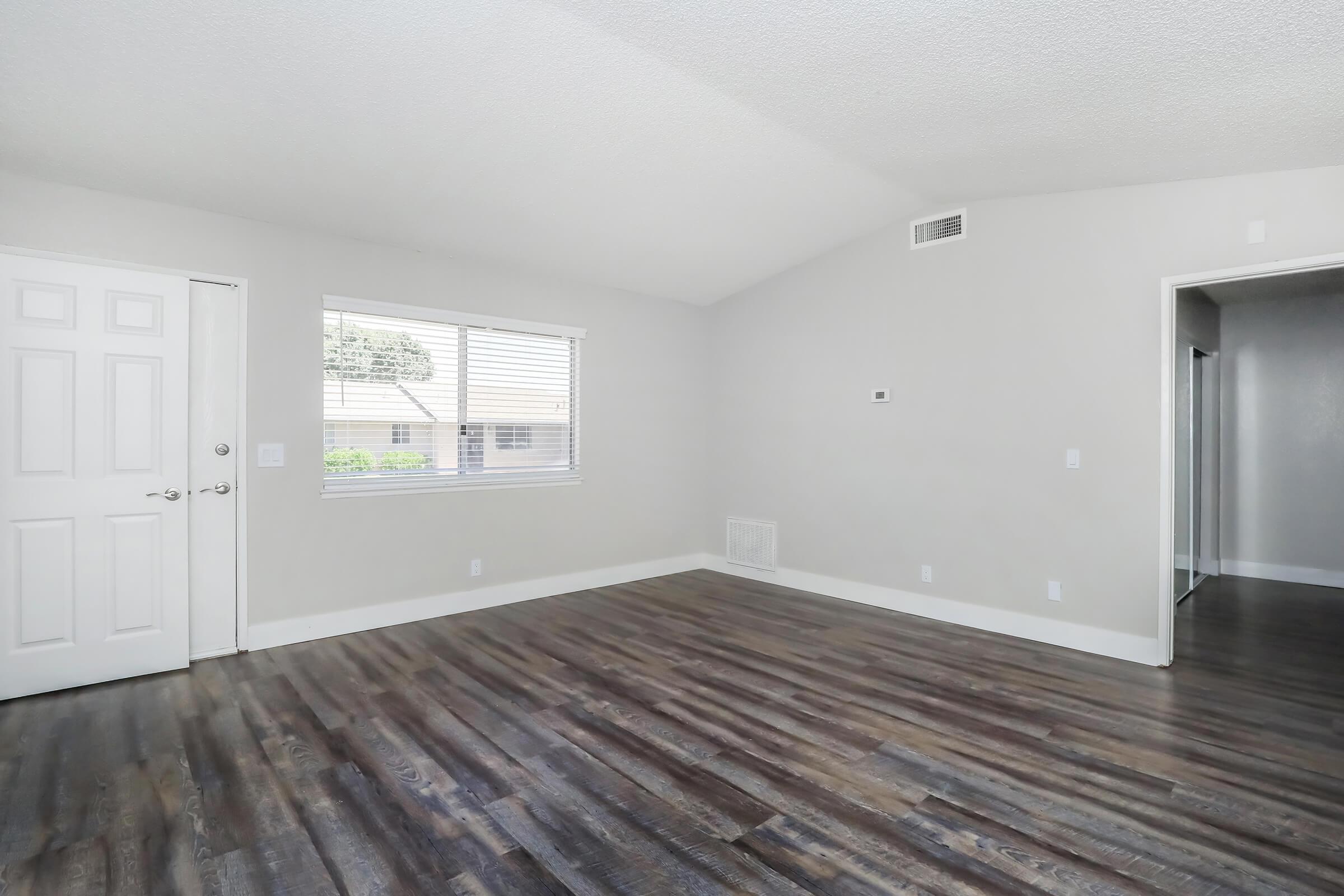 A spacious, empty room with a high ceiling and wooden floors. There is a door on the left leading outside, a large window allowing natural light in, and another door on the right leading to another area. The walls are painted in a light color, creating a bright and airy atmosphere.