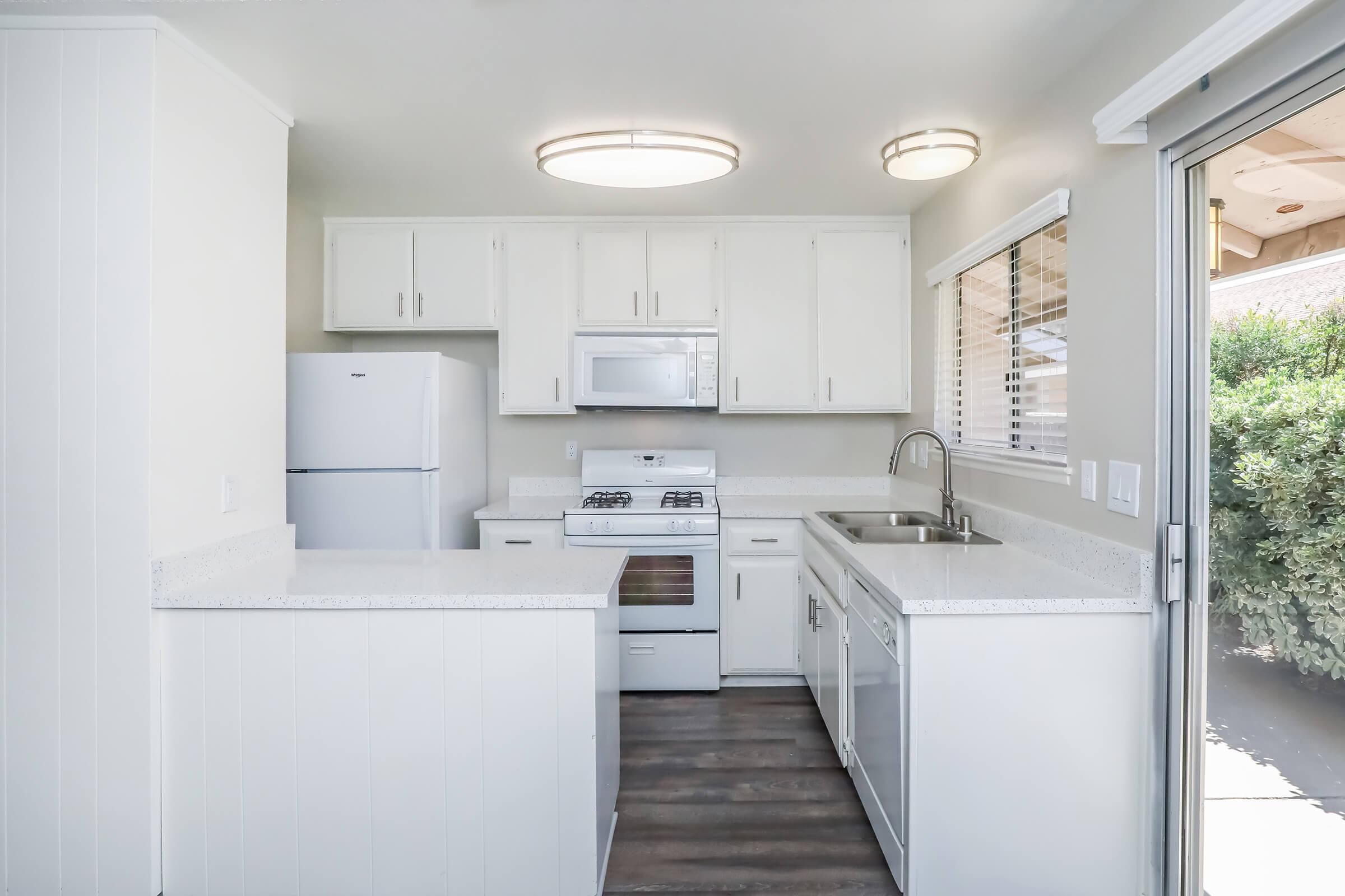 A modern kitchen featuring white cabinetry, a refrigerator, a gas stove, and a microwave. The countertop is light-colored with a sink, and there is a large window allowing natural light. The floor is dark wood, creating a contrast with the bright kitchen elements.