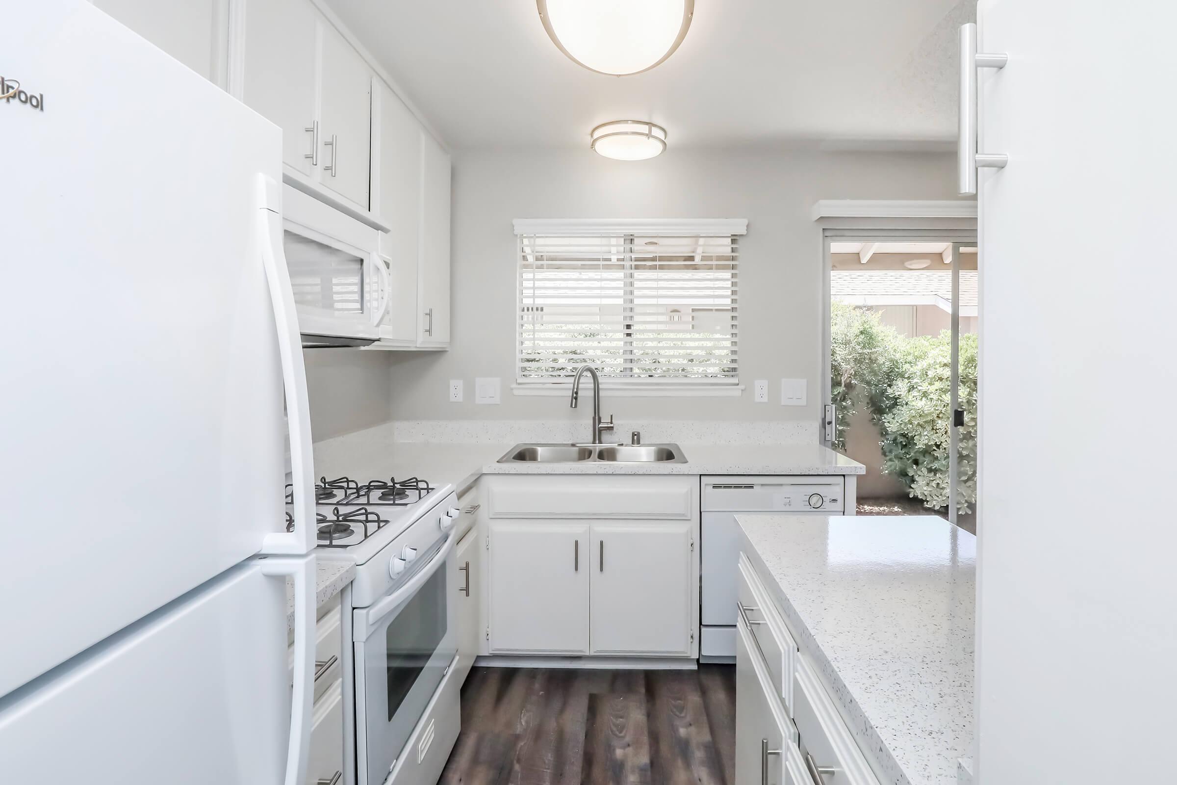 Modern kitchen with white cabinetry, a double sink, and stainless steel appliances, including an oven and dishwasher. There is a large window letting in natural light, and a small patio area visible through the door. The countertops are light-colored, and the floor features dark wood-like laminate.
