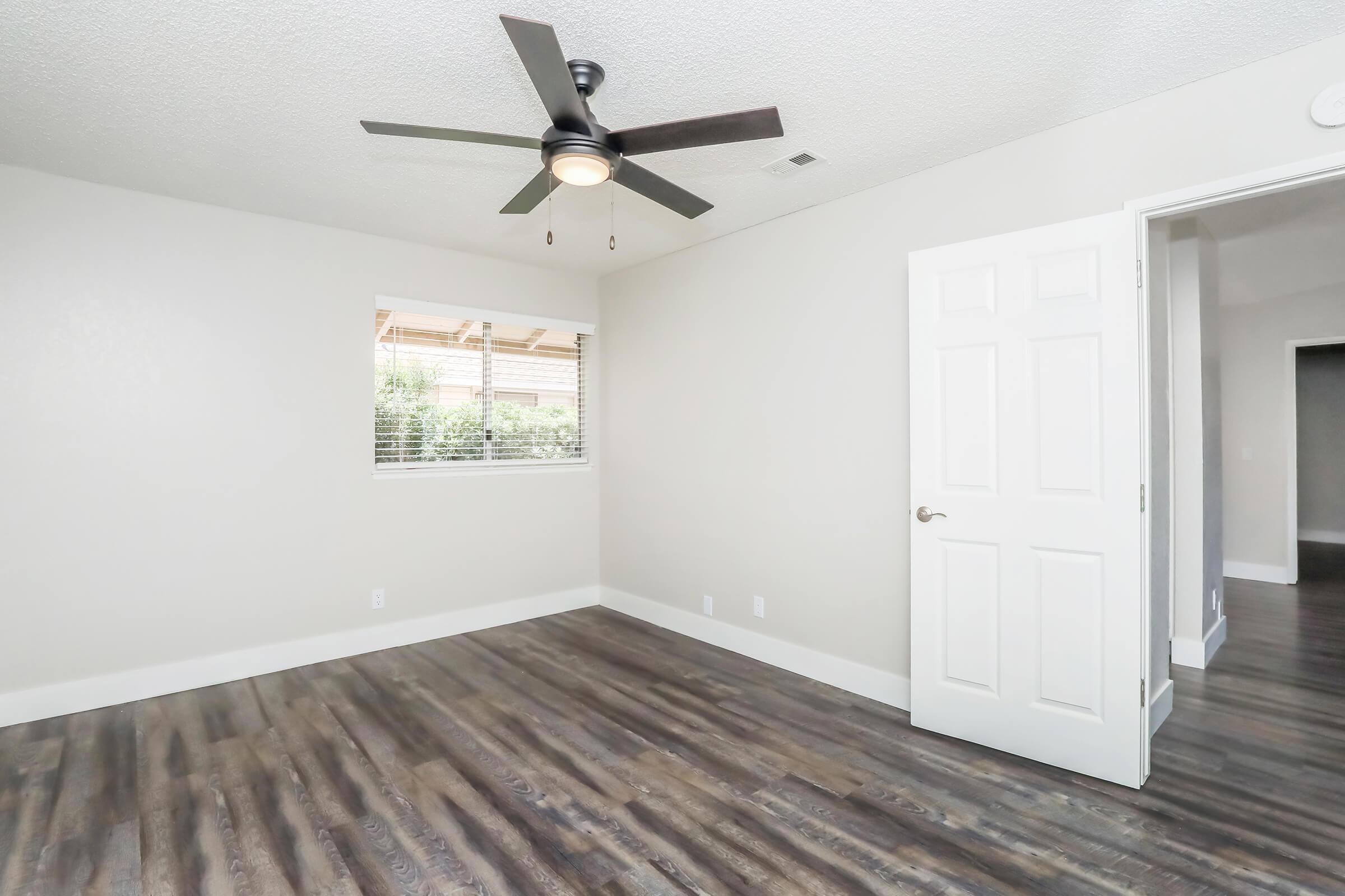 A well-lit empty room featuring light gray walls, a ceiling fan with a light fixture, and a window with blinds. The floor is made of dark wood laminate, and there is a white door partially open leading to another space. The room has a clean and modern aesthetic.