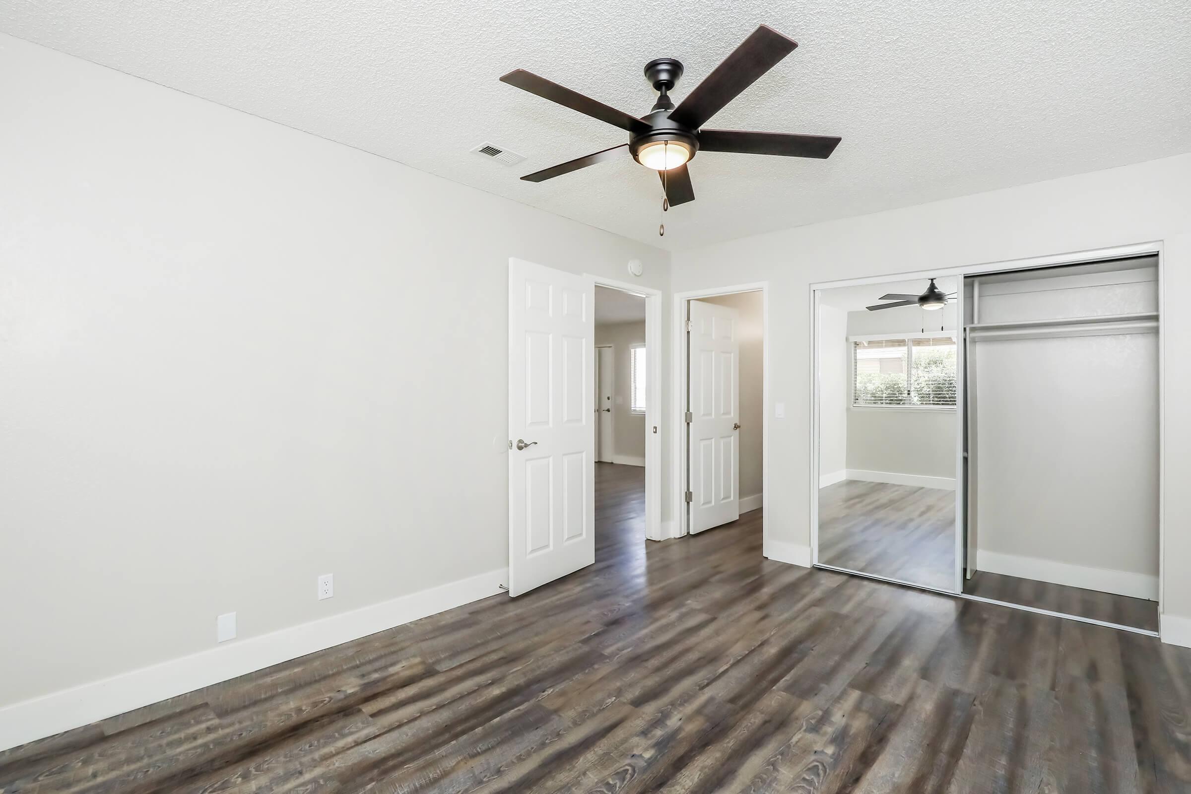 A spacious, well-lit room featuring a ceiling fan and light fixture, with two doors leading to adjacent areas. One door leads to a closet with sliding mirror doors, and the walls are painted a neutral color. The floor has a wood-like finish, adding warmth to the space. Windows let in natural light.
