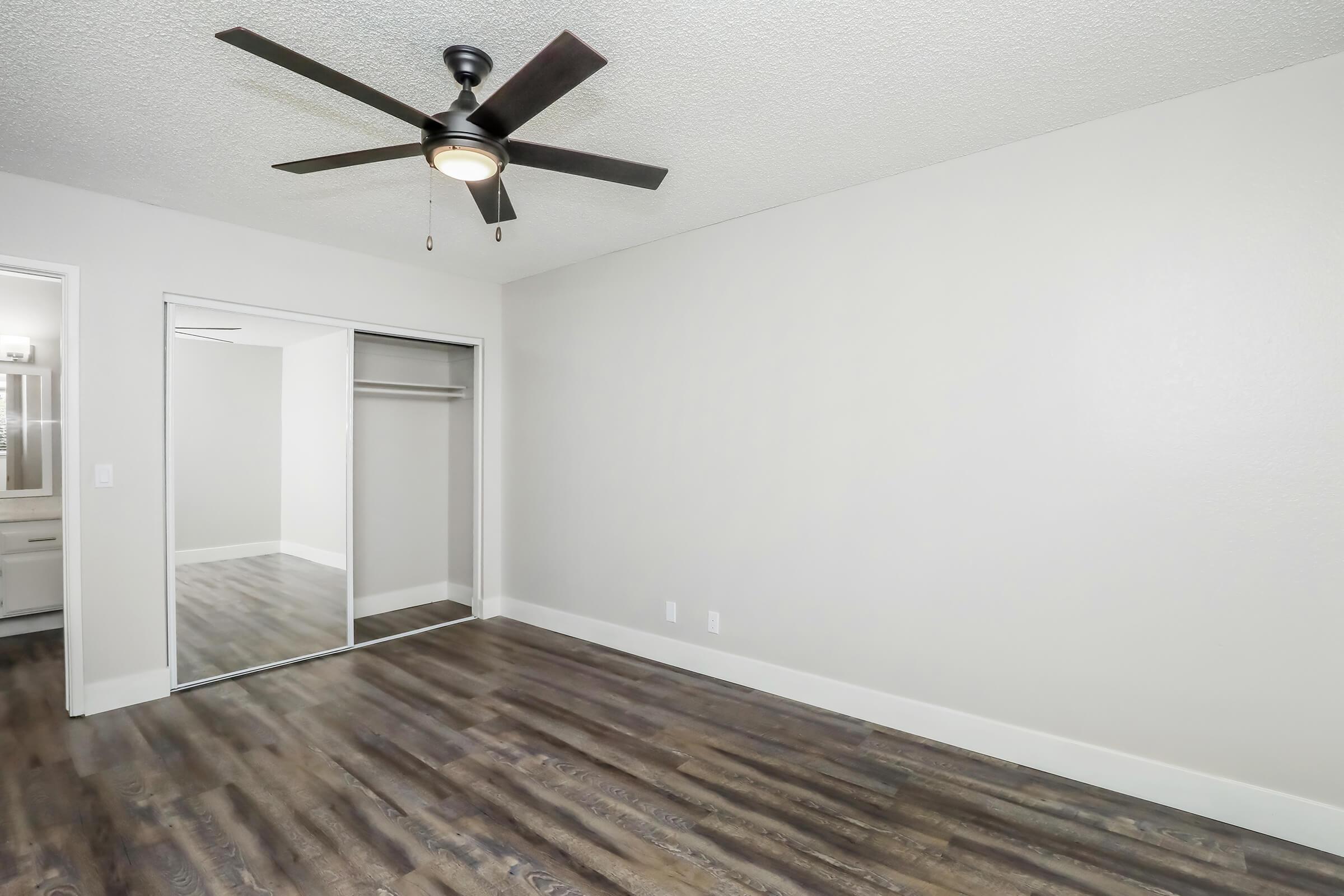 Room interior featuring a ceiling fan, hardwood-style flooring, and a mirrored closet door. The walls are painted in a light neutral color, creating a spacious feel. A doorway visible on the left leads to another area. The room is empty, highlighting its potential use as a bedroom or office space.