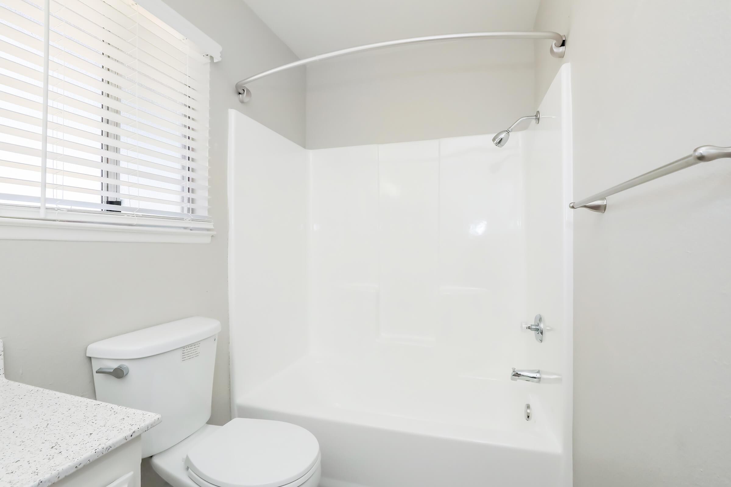 A clean bathroom featuring a white bathtub with an attached shower, a toilet, and a small window with white blinds allowing natural light. The walls are painted light, creating a bright and spacious atmosphere. A towel bar is visible next to the bathtub.
