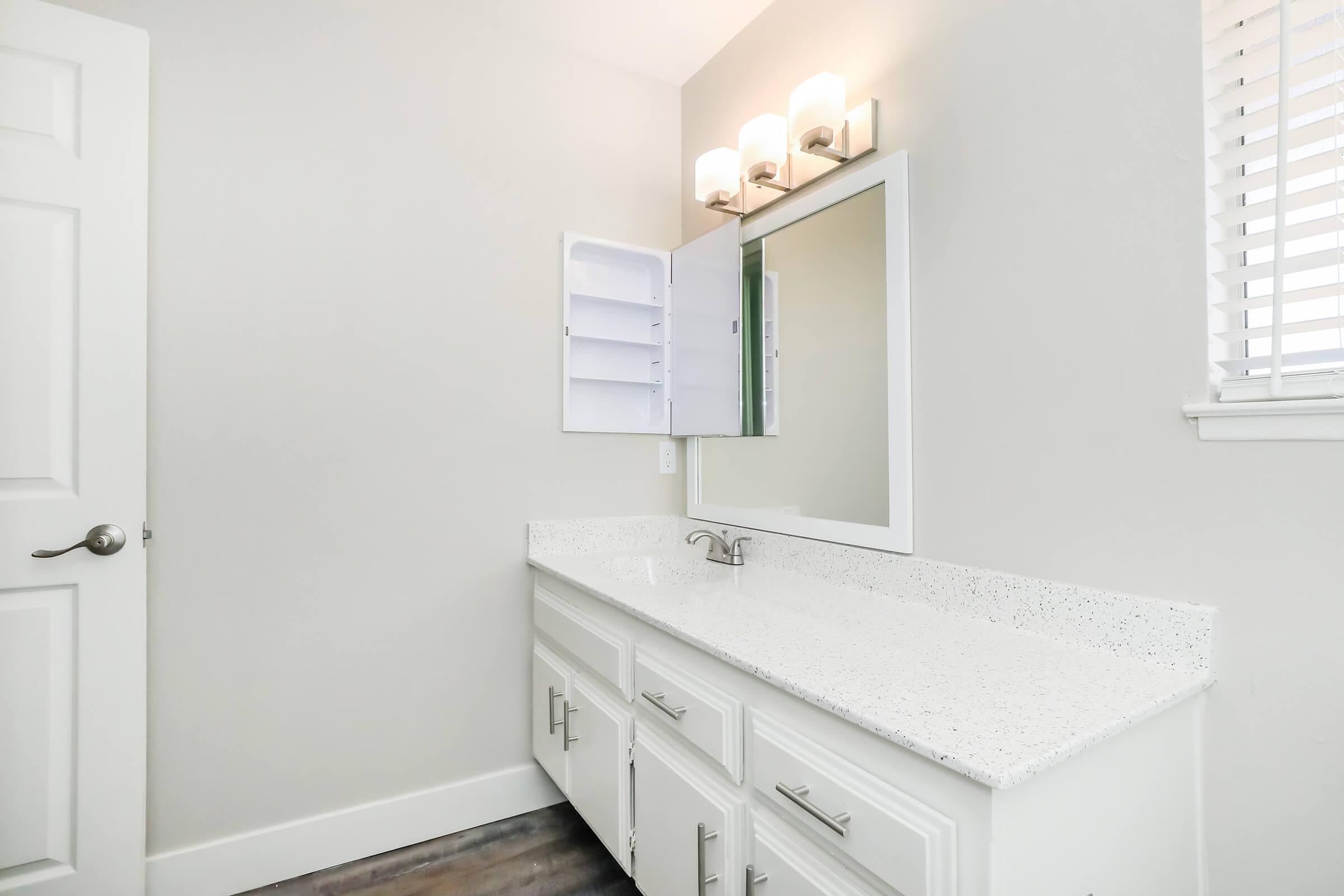 A modern bathroom with a white vanity featuring a speckled countertop. Above the sink, there is a rectangular mirror and a wall-mounted light fixture with three bulbs. The walls are light-colored, and there is a window with blinds on the right side, providing natural light. A door is visible on the left.