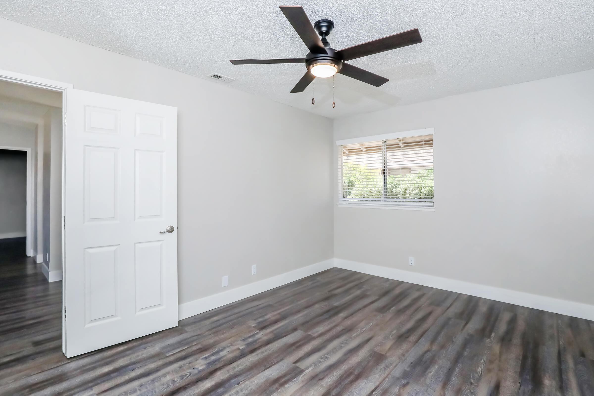 Empty room featuring a ceiling fan, hardwood-style flooring, and neutral-colored walls. A door leads to another space, while a window with blinds offers natural light. The overall ambiance is bright and airy, suitable for a bedroom or office.