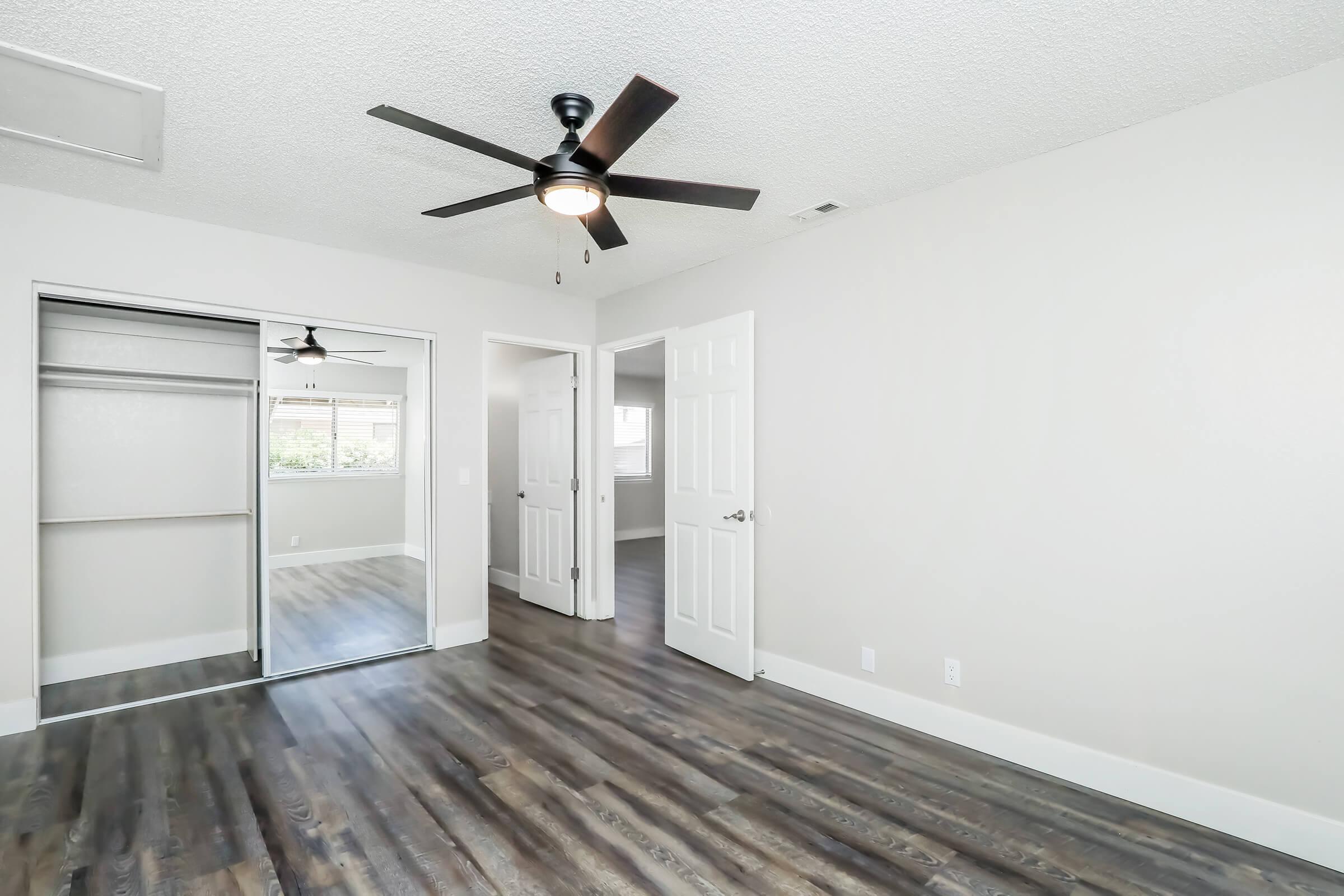 A well-lit bedroom featuring freshly painted light gray walls, a ceiling fan with light, and laminate flooring. The room includes a mirrored closet with sliding doors and white double doors leading to another space. Natural light enters through a window, enhancing the inviting atmosphere.