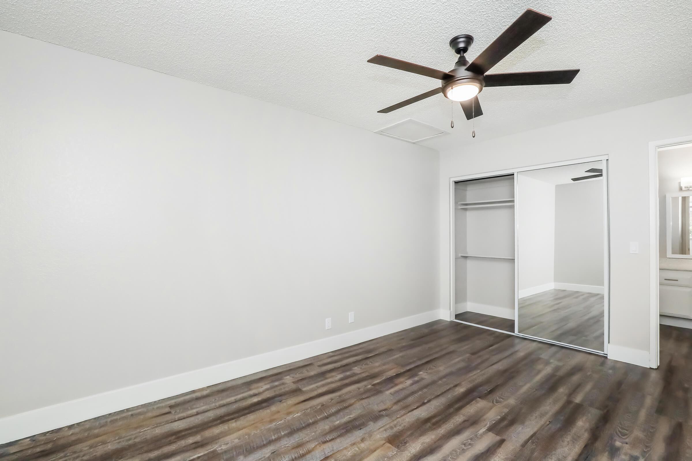 Empty bedroom with a ceiling fan, light-colored walls, and laminate flooring. There is a mirrored sliding closet door on one side, and the space is well-lit. The room has a clean, modern aesthetic, suitable for personal decoration or furnishing.