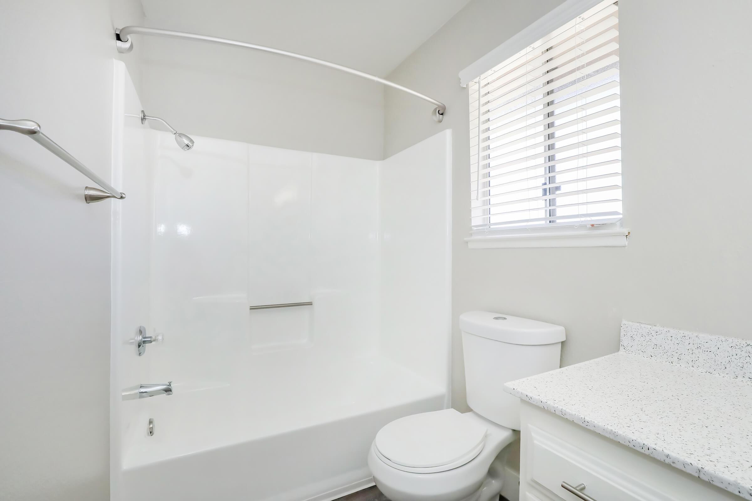 A clean, modern bathroom featuring a white bathtub and shower combination, a toilet, and a sink with a speckled countertop. Natural light enters through a window with white blinds. The walls are painted in a light color, enhancing the bright and spacious feel of the room.