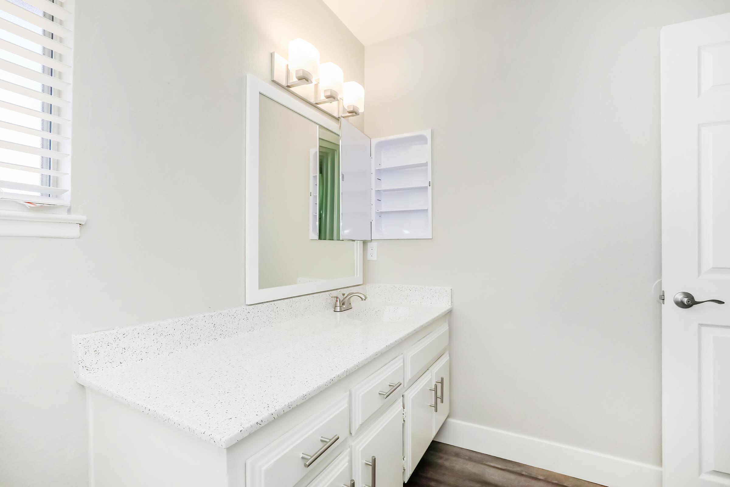 A modern bathroom featuring a white countertop with a sink, a large mirror, and stylish light fixtures above. There's an open cabinet with shelves on the wall, and the room has light gray walls and wooden flooring. A door is visible on the right side of the image.