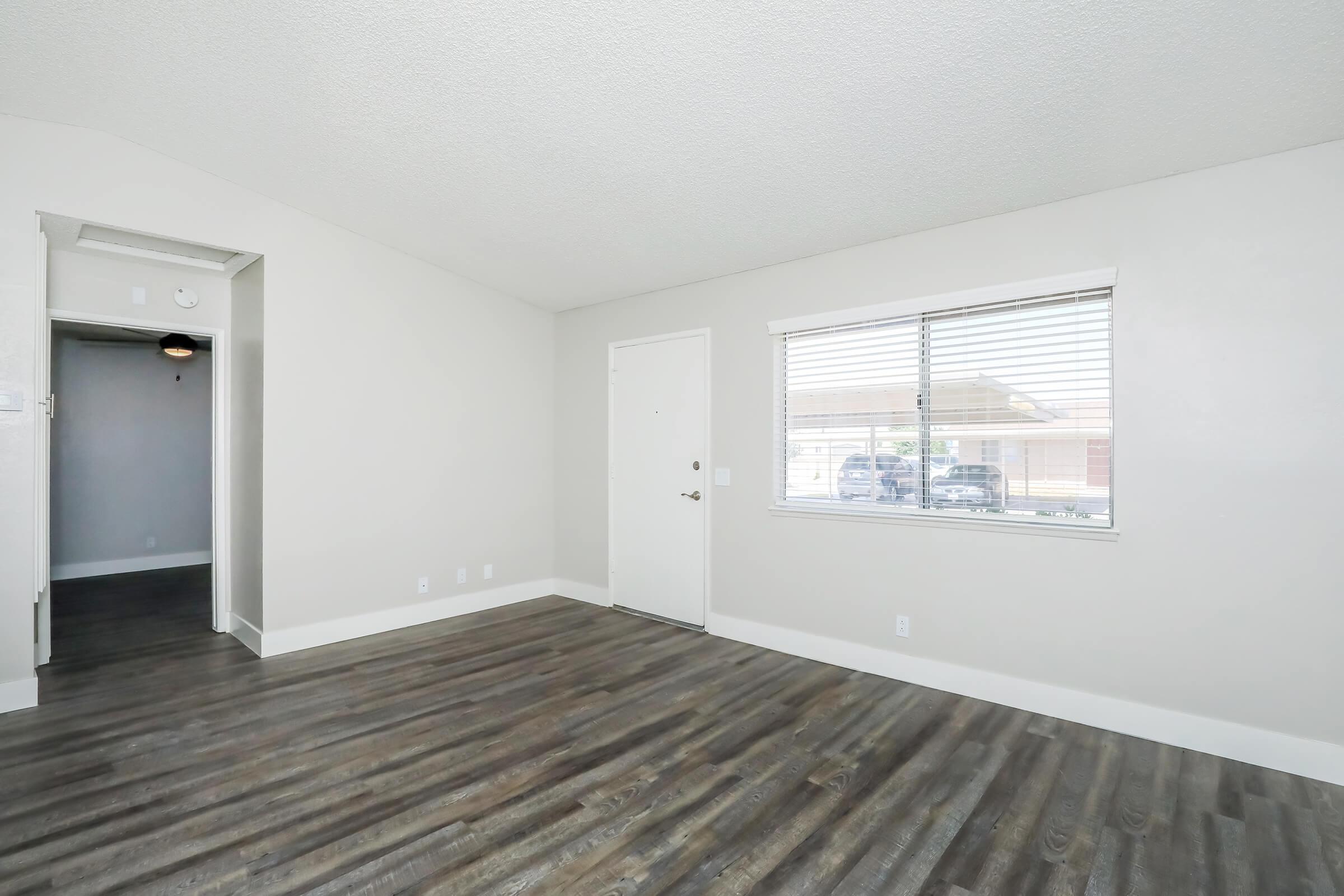 Spacious, minimalist living room with light-colored walls and dark wooden flooring. There is a large window allowing natural light, a front door, and an archway leading to another area of the house, creating an open and inviting atmosphere.