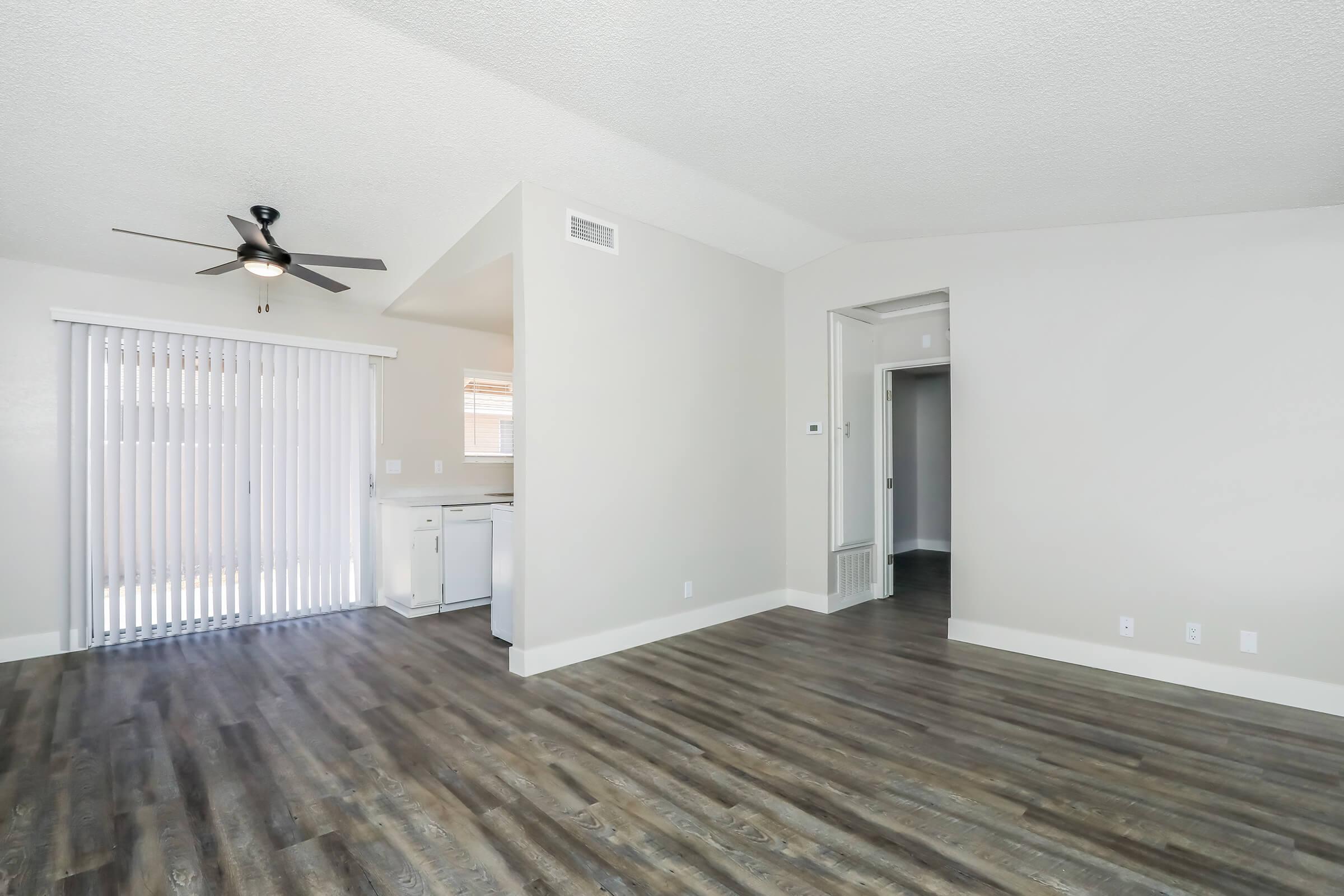 Spacious living area with wood-style flooring, a ceiling fan, and natural light from sliding glass doors. The adjacent kitchen is visible, showcasing a modern design. The walls are painted in a light color, enhancing the open feel of the room, and a doorway leads to a hallway.