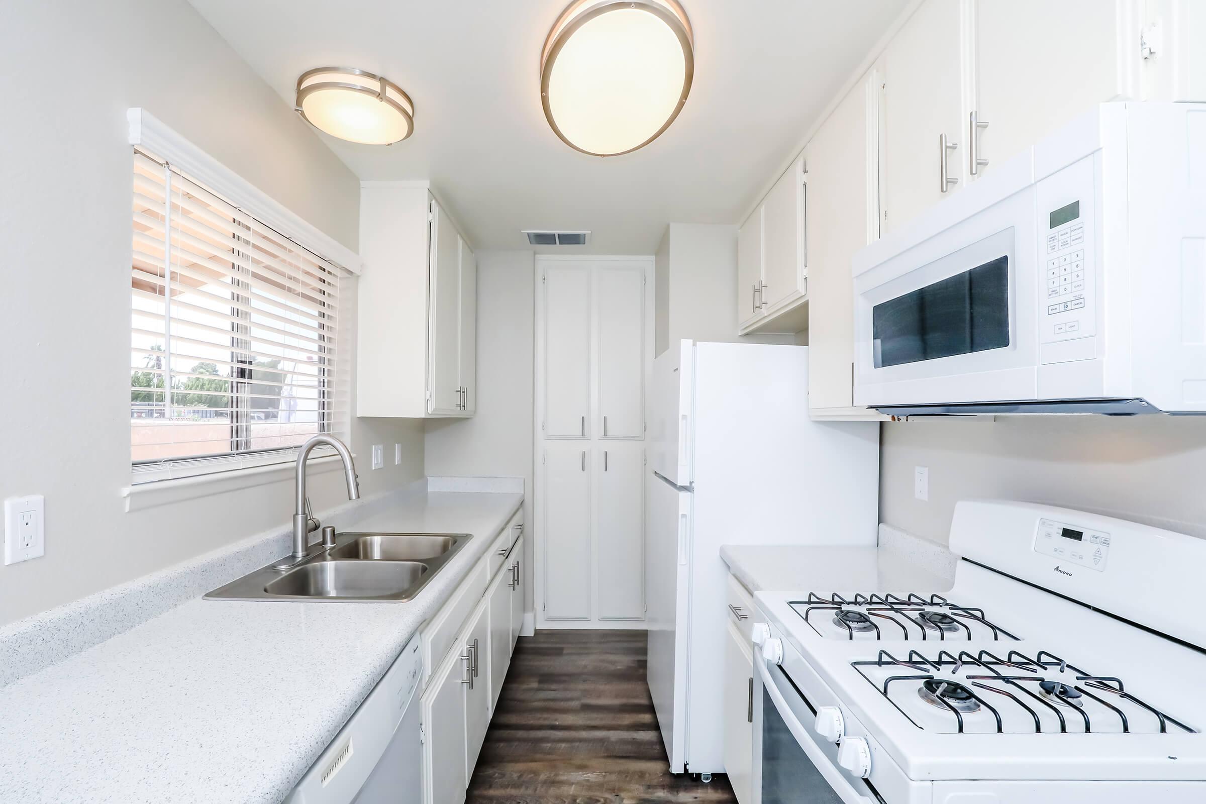 A modern kitchen featuring light-colored cabinets, a double sink, a white refrigerator, a microwave, and a gas stove. Natural light enters through a window with blinds, illuminating the countertop and appliances. The flooring is a dark wood, providing contrast to the overall bright aesthetic.