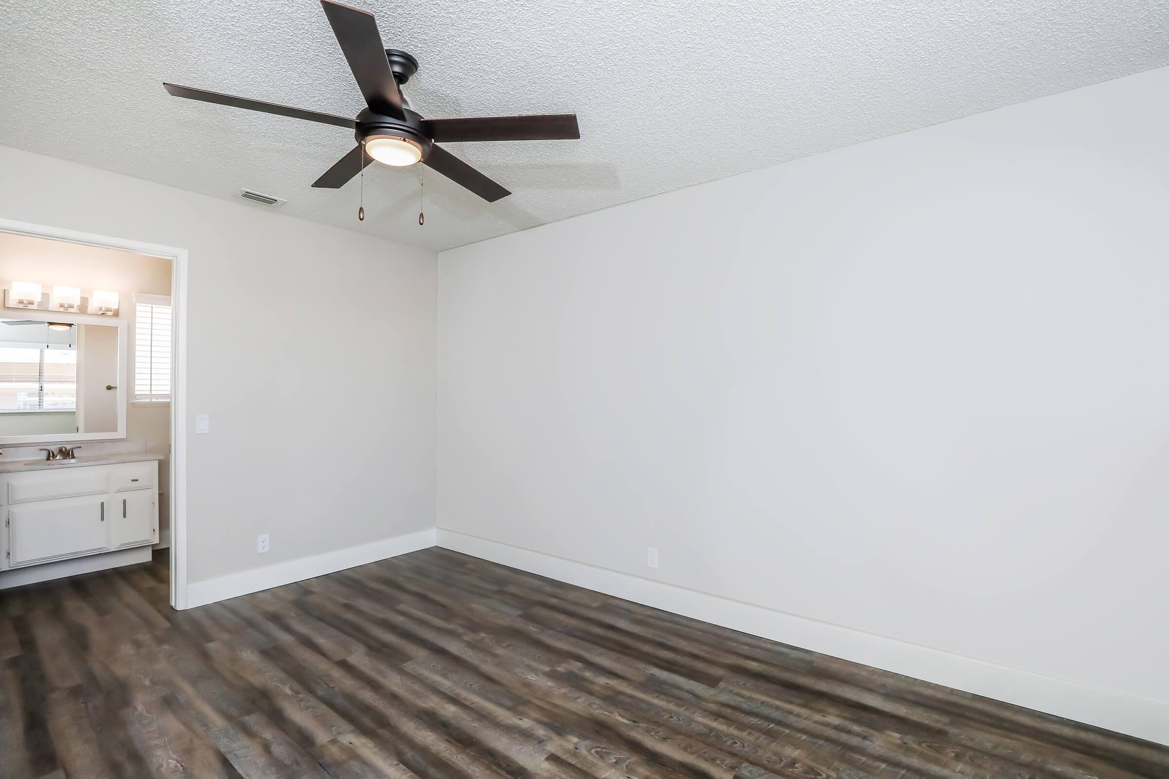 A spacious, empty room with light gray walls and a ceiling fan. Wood-like flooring runs throughout the space. A doorway leads to a bathroom area, visible in the background, featuring a vanity with a mirror and bright lighting. The room is well-lit and has a clean, modern aesthetic.