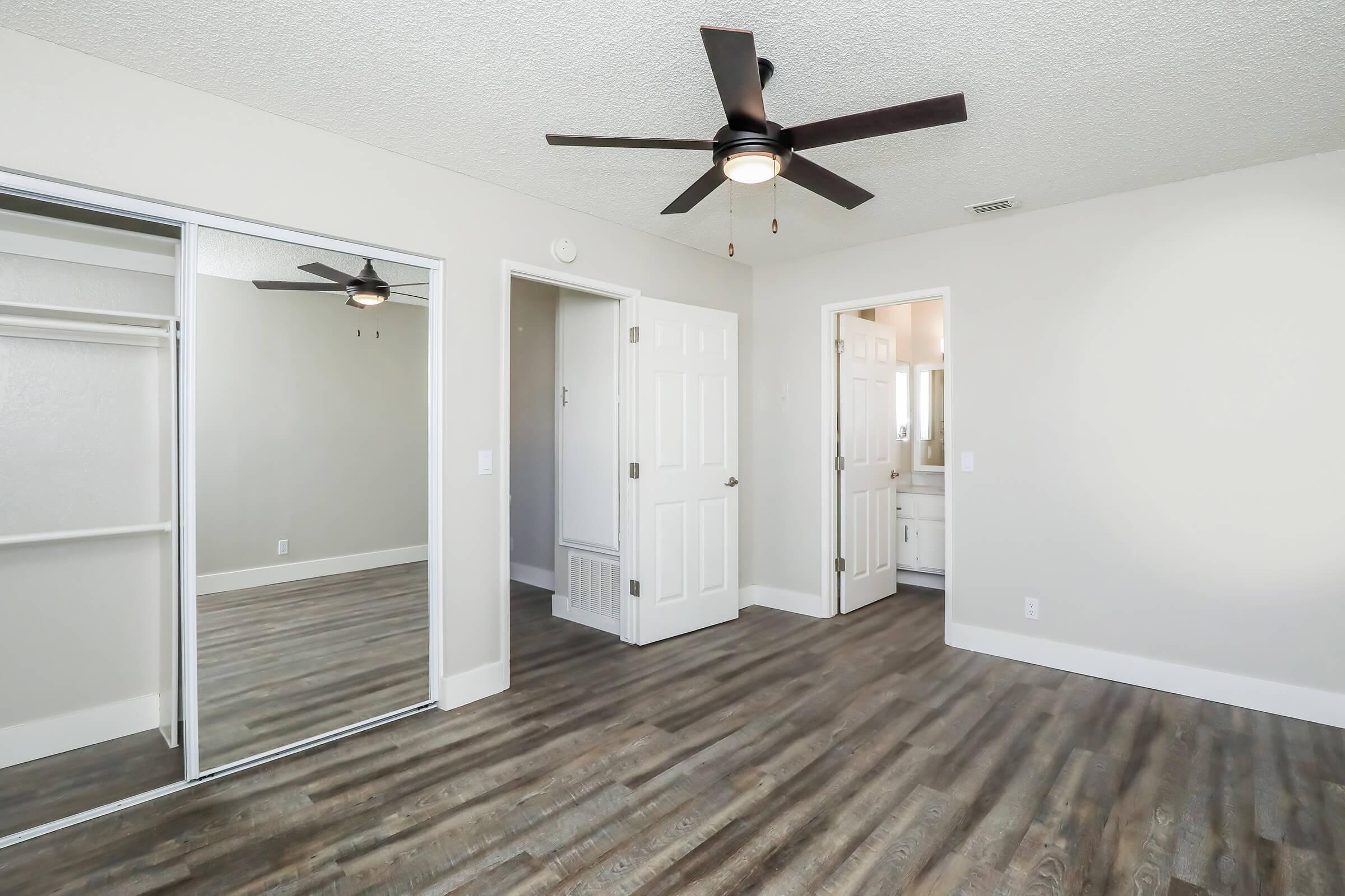 A spacious bedroom featuring light-colored walls, a ceiling fan, and a mirrored closet. The room has wooden laminate flooring and an open doorway leading to a bathroom area. Natural light brightens the space, creating a warm and inviting atmosphere.