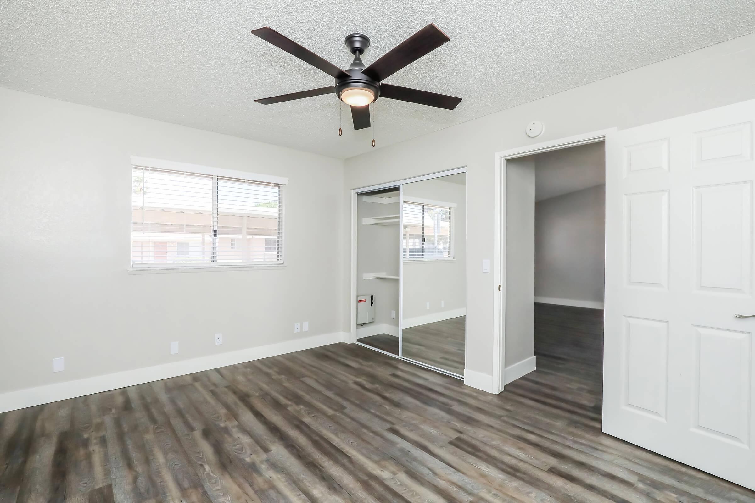 Spacious empty room featuring light-colored walls, a ceiling fan, and a large mirror-fronted closet. The floor is made of laminate wood, and there are windows with blinds letting in natural light. A doorway leads to another room in the background.