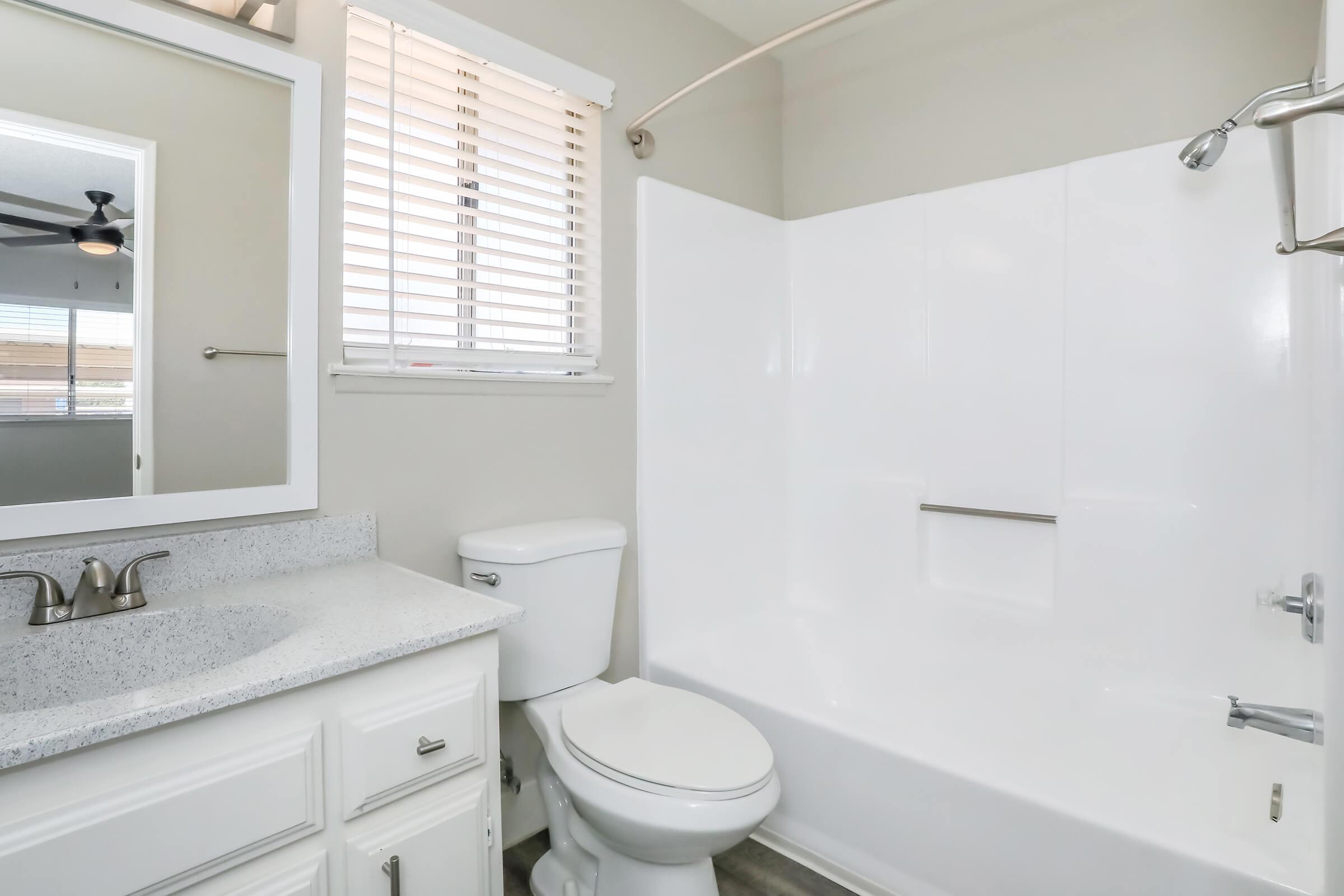 A clean, modern bathroom featuring a white bathtub with a shower, a toilet, and a sink with a granite countertop. The walls are painted light grey, and there's a mirror above the sink. Natural light comes through a window with blinds.