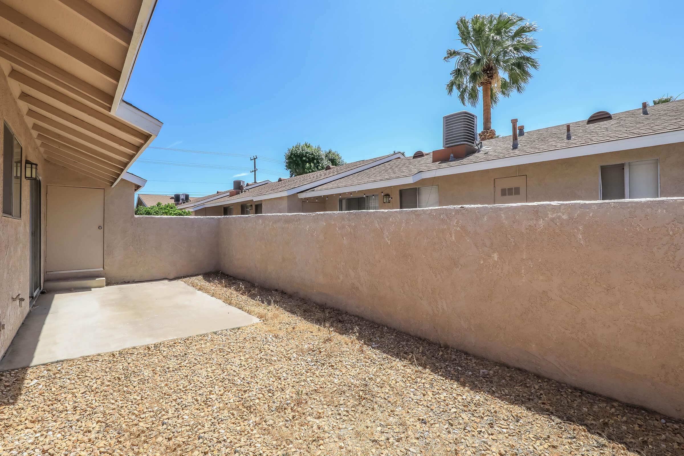 A narrow outdoor space with a concrete area and gravel, surrounded by a low wall. In the background, there are a few houses and a tall palm tree against a clear blue sky. Sunlight brightens the area, creating a warm and inviting atmosphere.