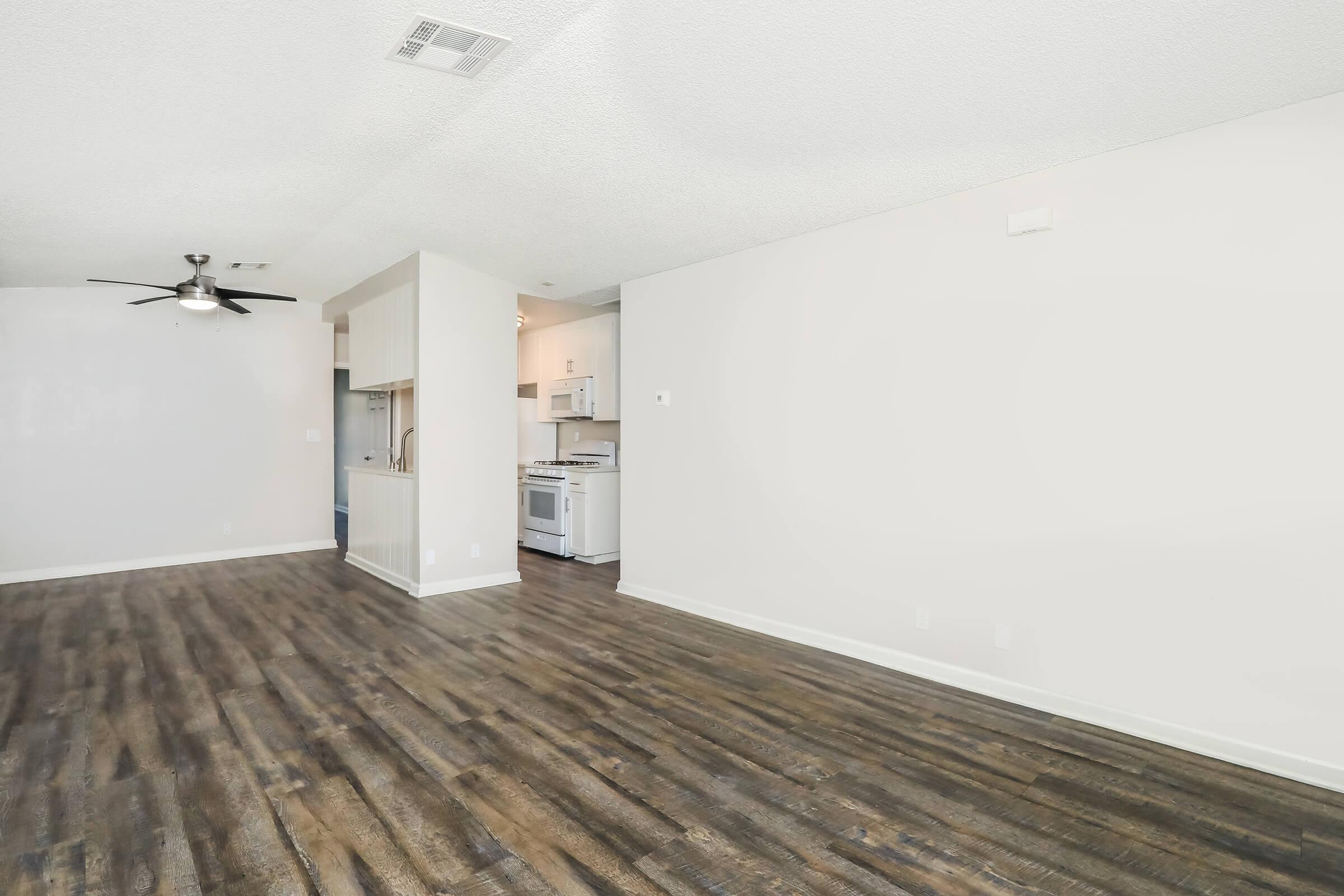 A spacious living area featuring a ceiling fan, light-colored walls, and hardwood-style flooring. An adjacent kitchen area is visible with white cabinetry and appliances, creating an open-concept layout that enhances the room's brightness.