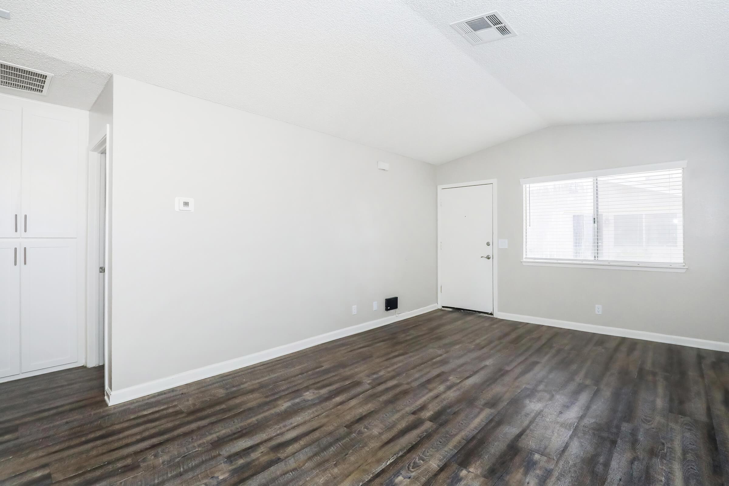 A spacious, empty living room with a high ceiling, light-colored walls, and dark wooden flooring. There's a white door leading outside and a window allowing natural light to enter. Built-in cabinets are visible on one side, adding to the room's modern and airy feel.