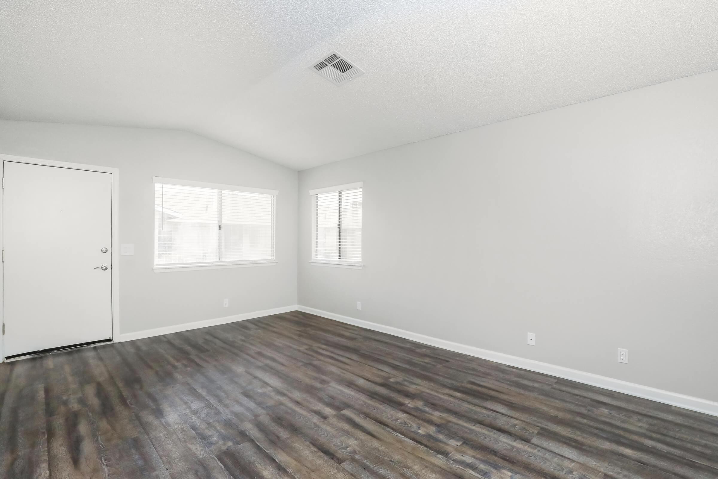 A spacious and empty living room with light gray walls, large windows allowing natural light, and a modern wood-like floor. The entrance door is visible on the left side of the image, contributing to a clean and minimalistic aesthetic.