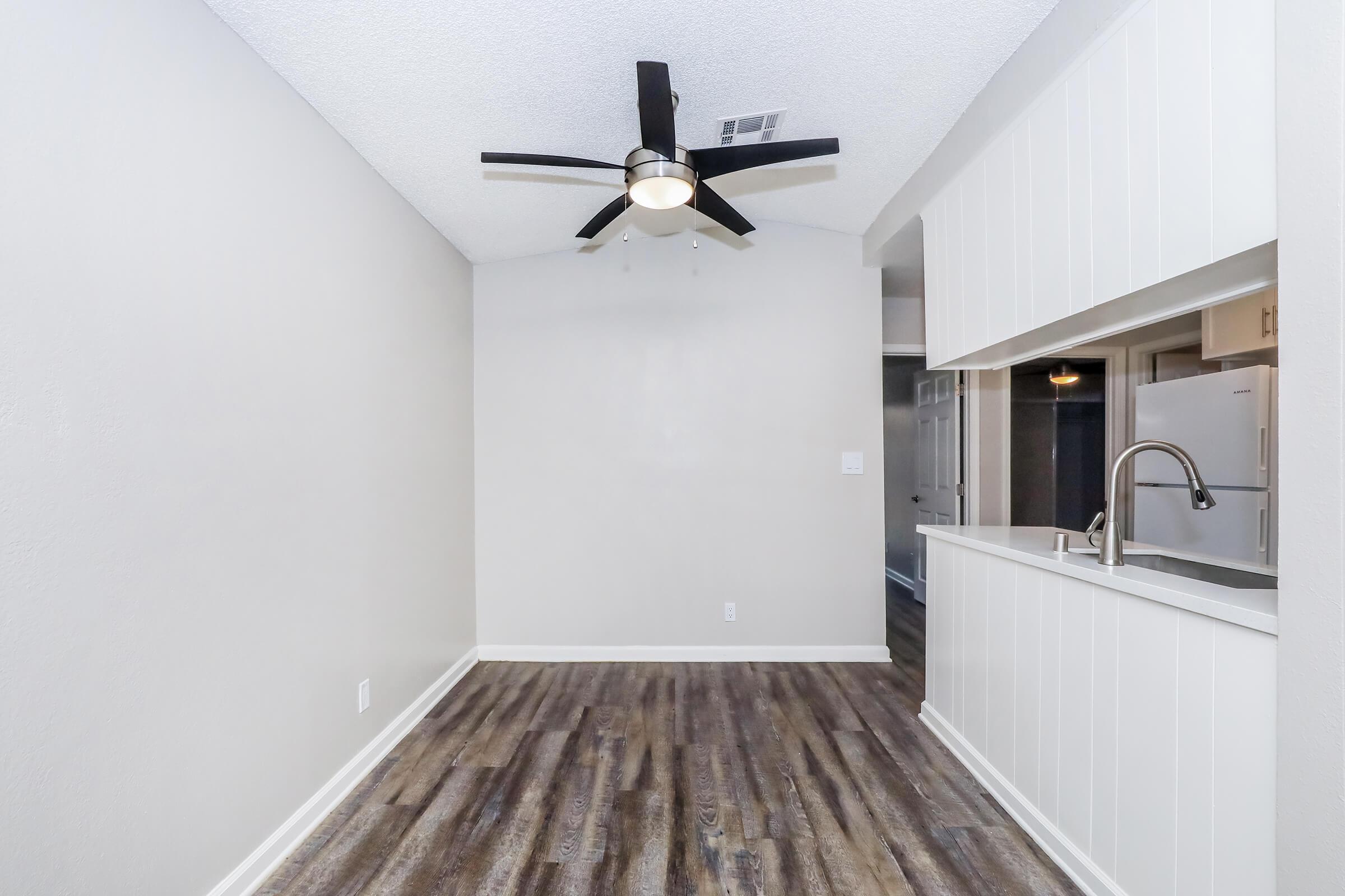 A modern living space featuring a light gray wall, a ceiling fan with black blades, and wood-style flooring. The room includes an open kitchen area with a bar overlooking the living space, and a doorway leading to another room. The overall design is clean and contemporary.