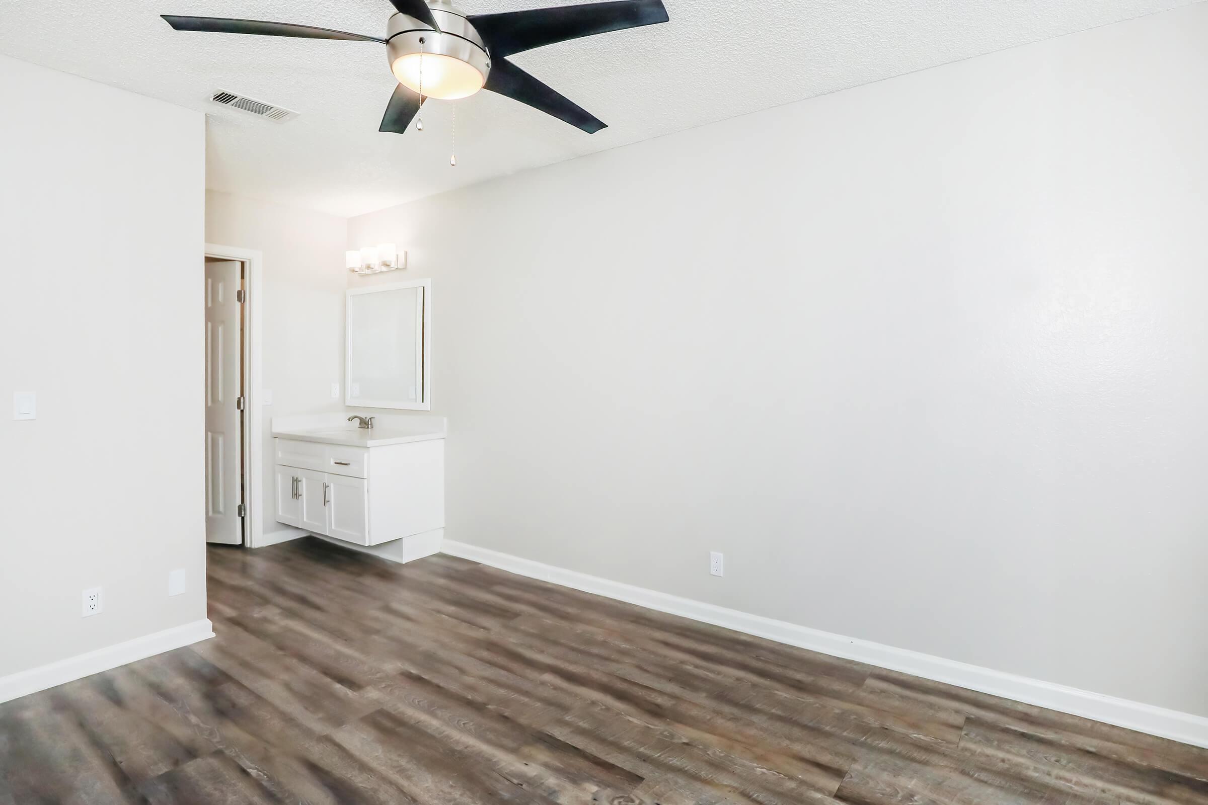 A spacious, well-lit room with light gray walls, a ceiling fan, and laminate flooring. To the left, there is a vanity with a mirror, and a doorway leading to another area. The overall atmosphere is clean and modern, suitable for a bedroom or a living space.