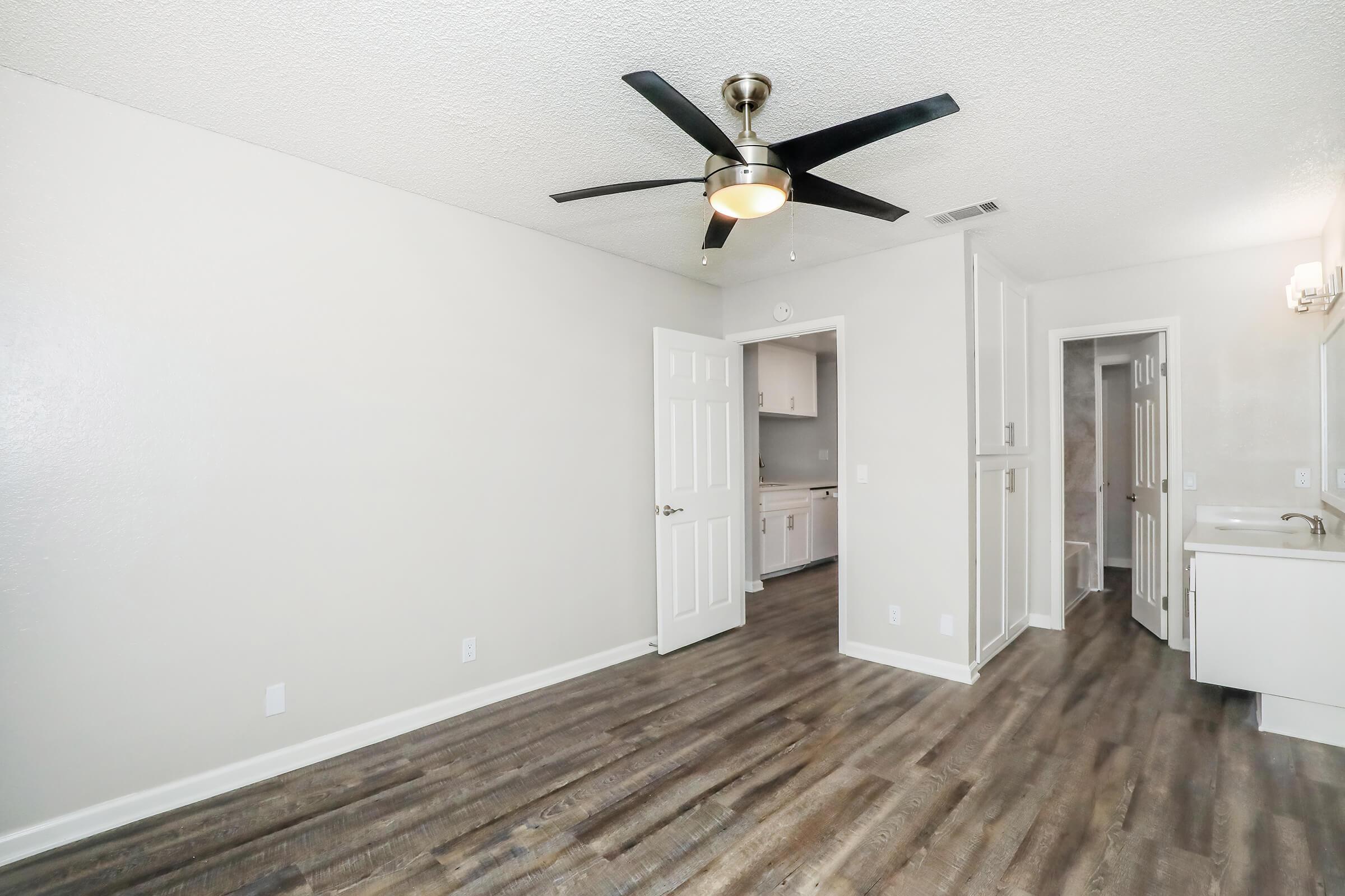 A spacious, light-colored living area with a ceiling fan and modern laminate flooring. The room features a doorway leading to a kitchen and a bathroom area, with neutral walls and ample natural light.