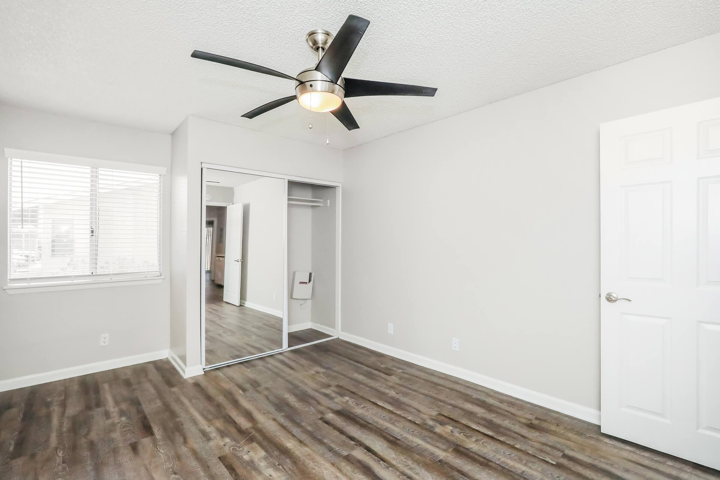 A bright, empty bedroom featuring light gray walls, a ceiling fan with a modern design, and a large mirrored closet. The floor is a warm-toned laminate, and there's a window with blinds allowing natural light to enter the room. The space is clean and ready for furnishing.