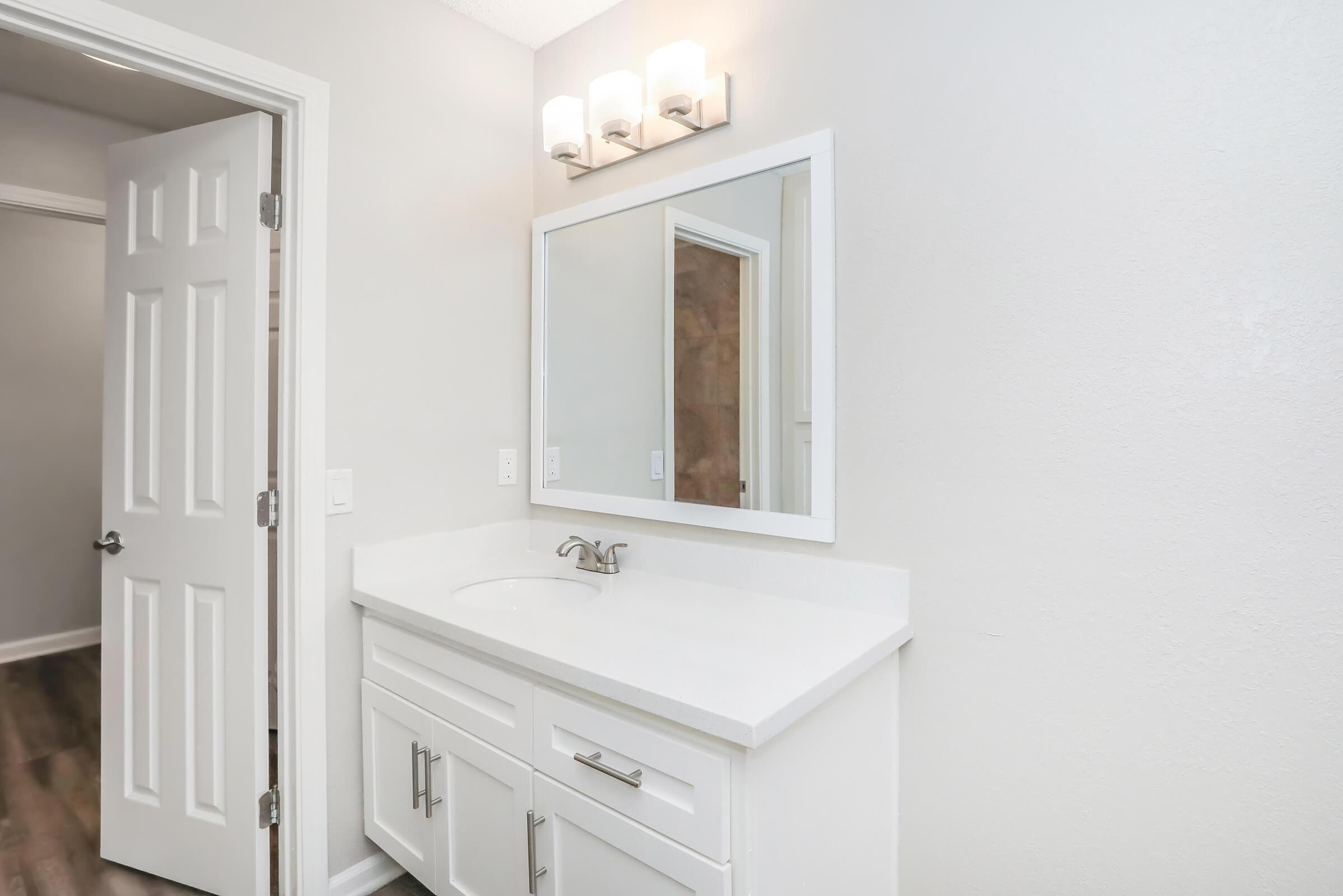 A modern bathroom vanity featuring a white countertop, a large mirror, and wall-mounted light fixtures. The light gray walls enhance the spacious feel, and a closed door is visible on the left. The flooring is a dark wood-like laminate, adding contrast to the overall design.