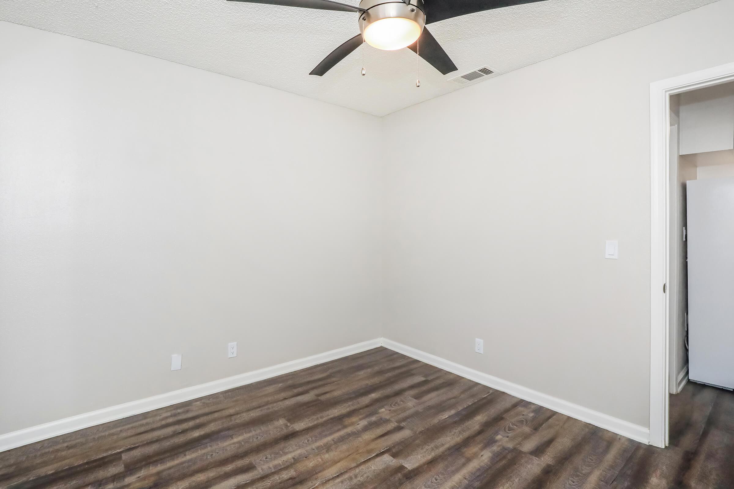 Empty room featuring light gray walls and a dark wood floor. A ceiling fan is visible, and there is a doorway leading to another area. The space has no furniture, creating a minimalist and open feel.