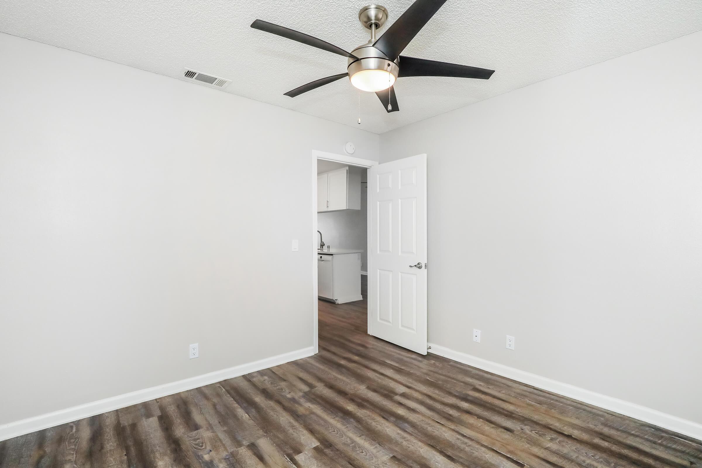 An empty room featuring a light-colored wall with a ceiling fan. A doorway leads to another area, possibly a kitchen, visible in the background. The floor is finished with wood-like laminate, providing a modern and spacious appearance.