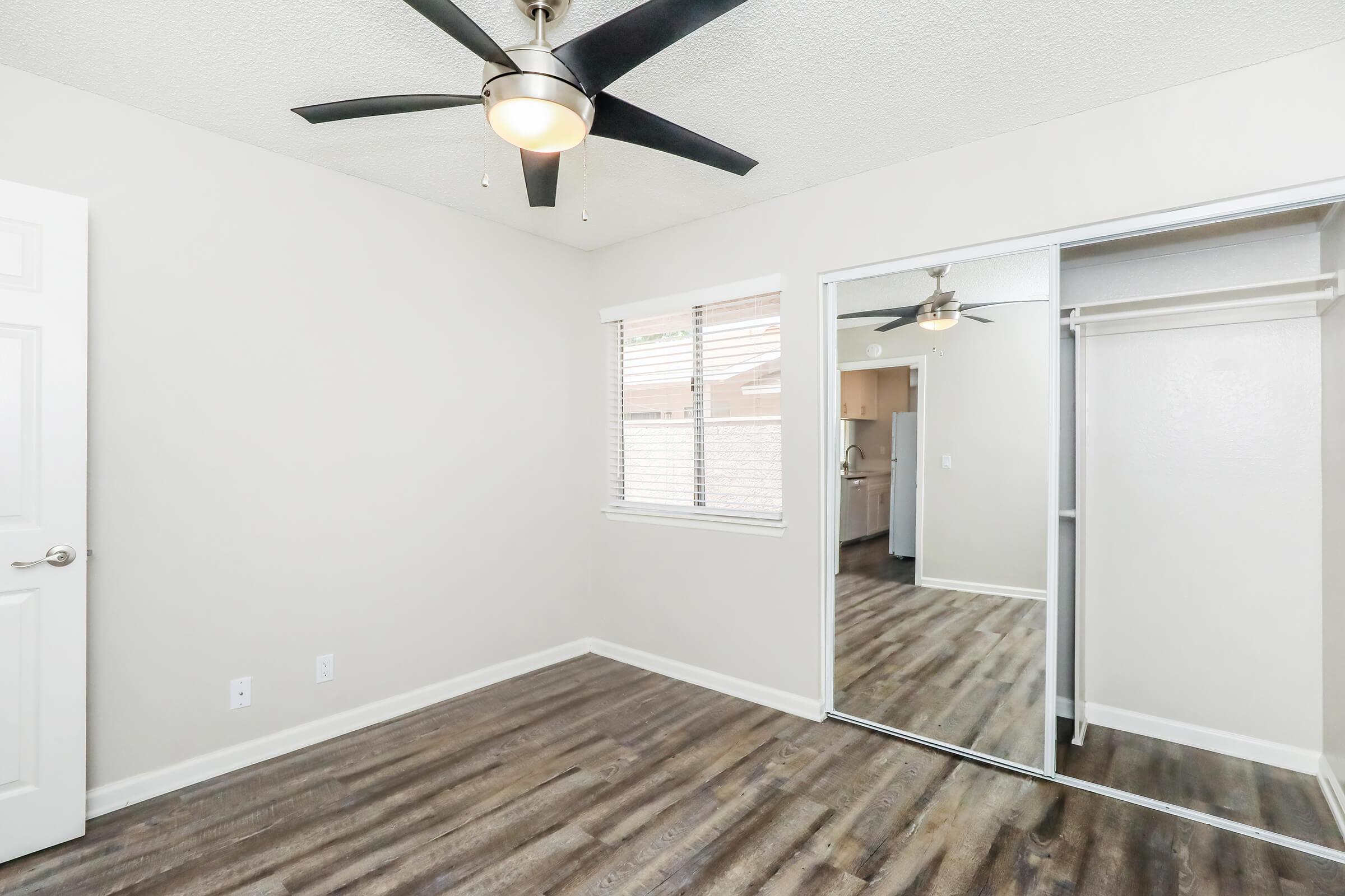 A well-lit room featuring a ceiling fan, neutral-colored walls, and a large mirror closet. The floor has a wood-like finish, and there are windows allowing natural light. The space appears clean and modern with minimal furnishings.