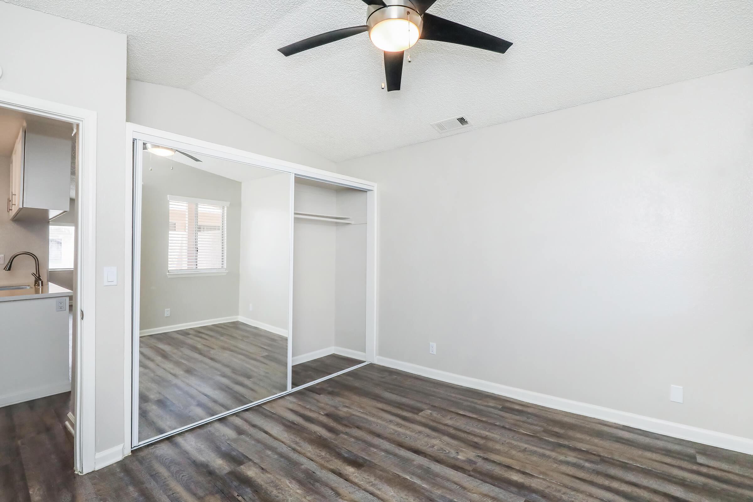 A spacious room with a ceiling fan and large mirrored closet doors. The walls are painted in a light color, and there are windows letting in natural light. The flooring is wood-like laminate, creating a modern and inviting atmosphere.