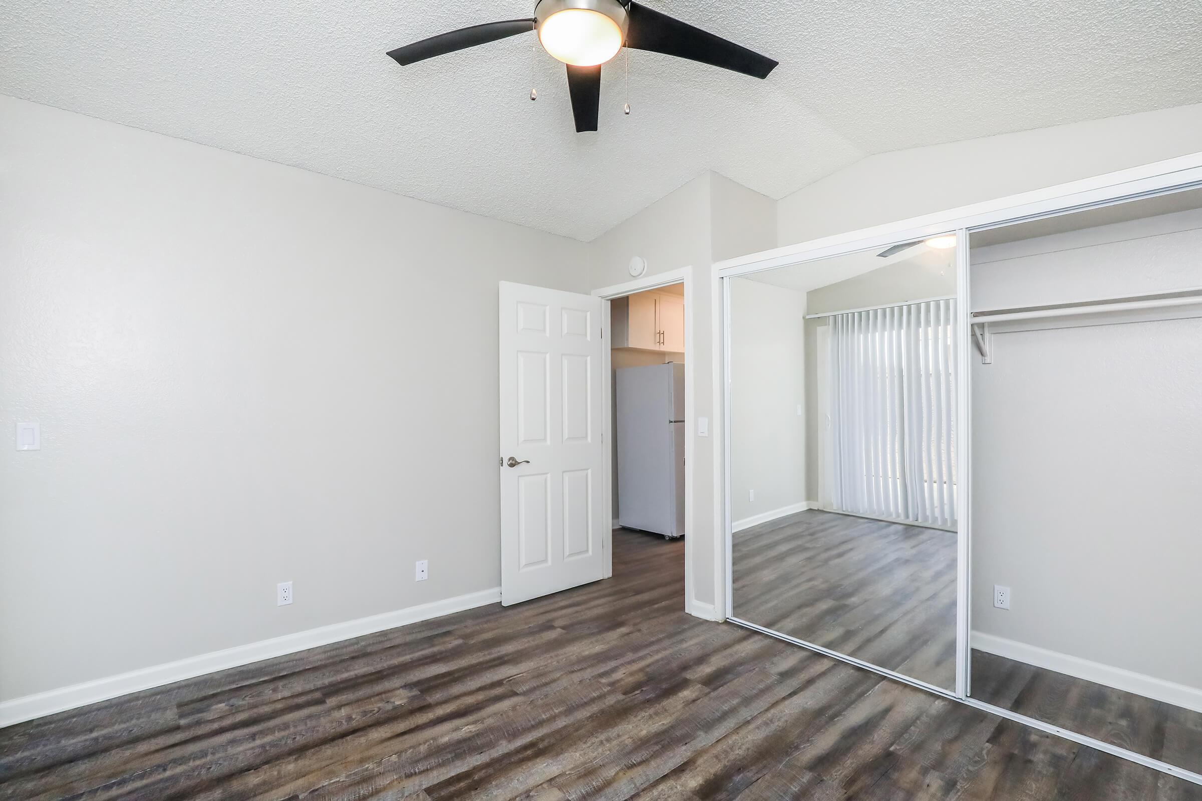Interior view of a room featuring light-colored walls, a ceiling fan, and hardwood-like flooring. A mirrored closet door is visible, reflecting the room's light. A doorway leads to another area, while behind it, a glimpse of a space with light coming through vertical blinds can be seen.