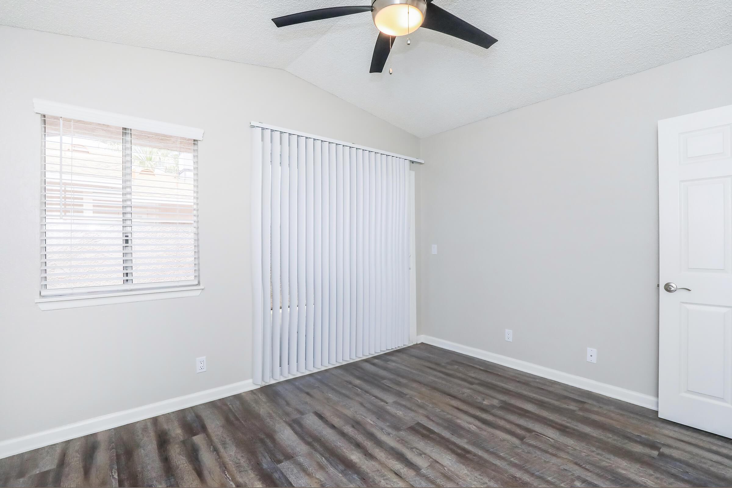 A well-lit interior room featuring a ceiling fan, light-colored walls, and a large window with blinds. The floor is covered in wood-like laminate, and there are white vertical blinds covering the window. The space is empty, providing a clean and minimalist look.