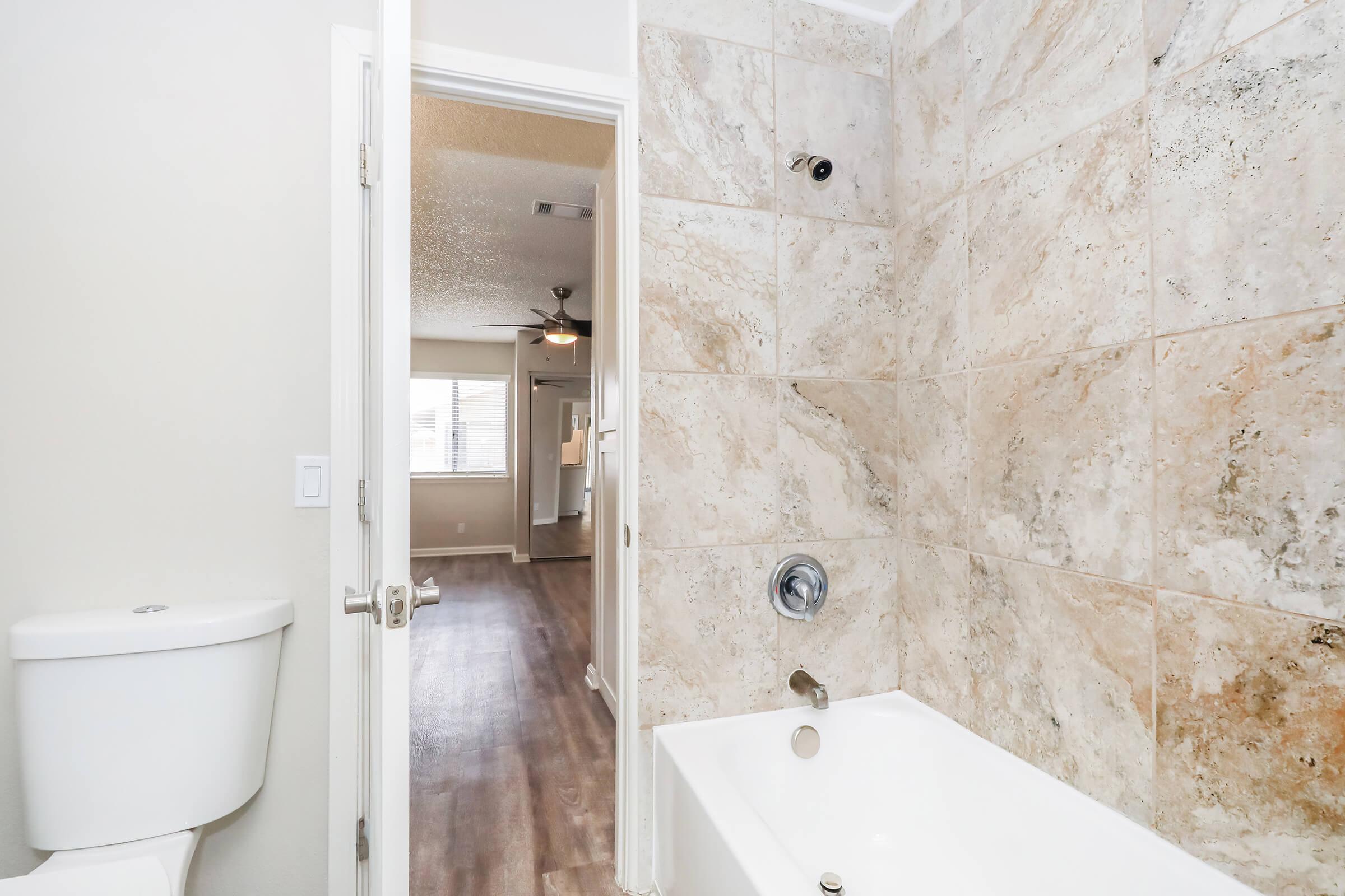 A modern bathroom featuring a white toilet and a bathtub with a sleek faucet. The wall is adorned with large, textured tiles. In the background, a doorway leads to a well-lit room with a ceiling fan and windows, indicating an open space beyond the bathroom.