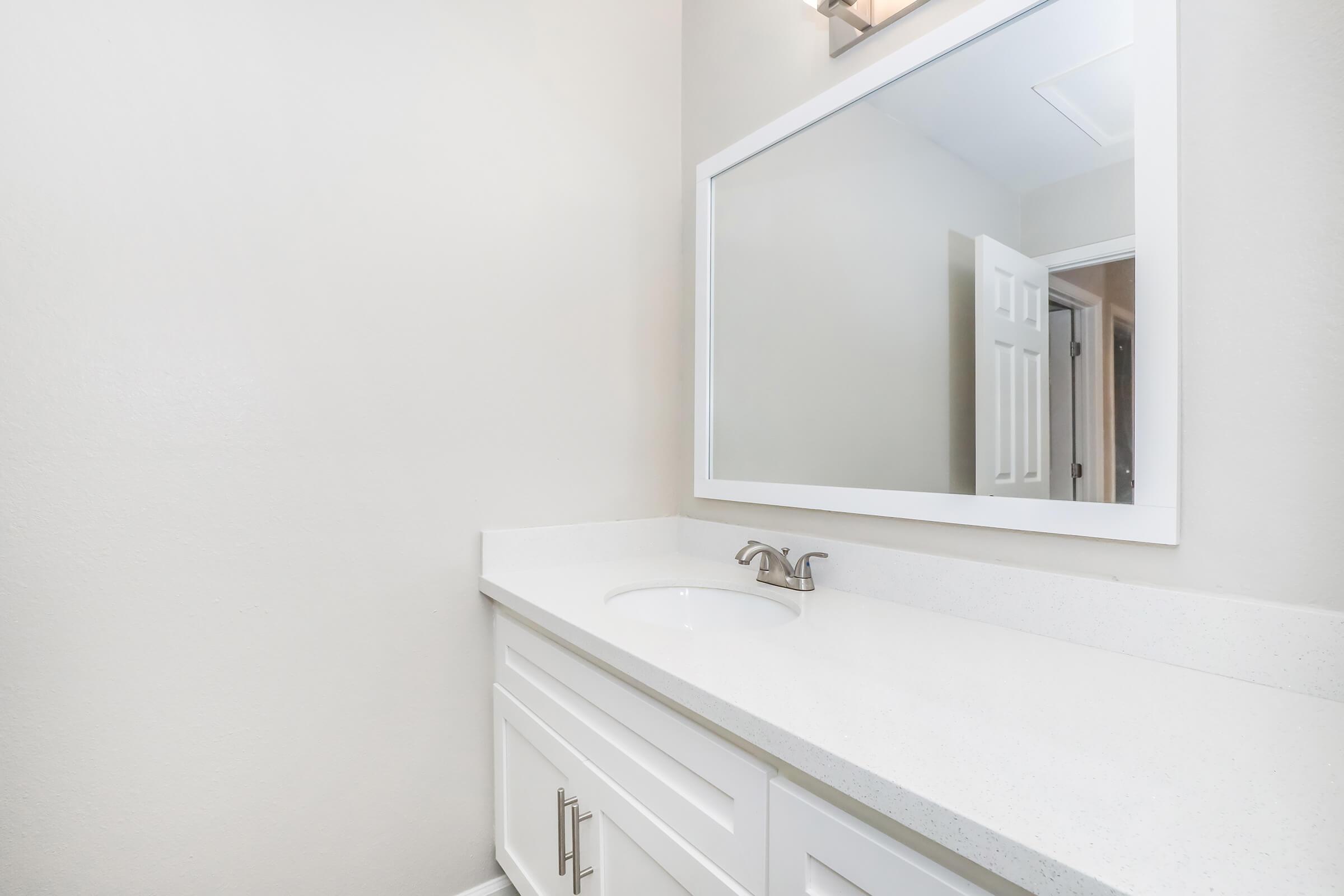 A modern bathroom featuring a clean white vanity with a sink, a large rectangular mirror above it, and neutral-colored walls. A door is visible in the background, adding depth to the space. The overall design is bright and minimalist.