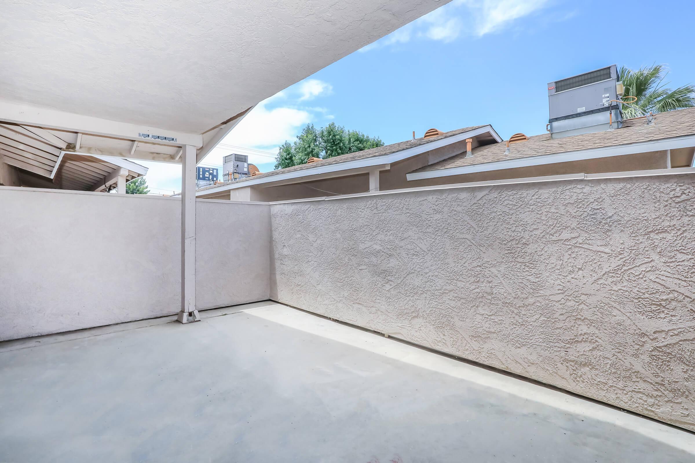 A spacious outdoor balcony area with a textured wall and a concrete floor. The sky is bright with a few clouds, and there are rooftops and air conditioning units visible in the background. The setting gives a sense of openness and sunlight.