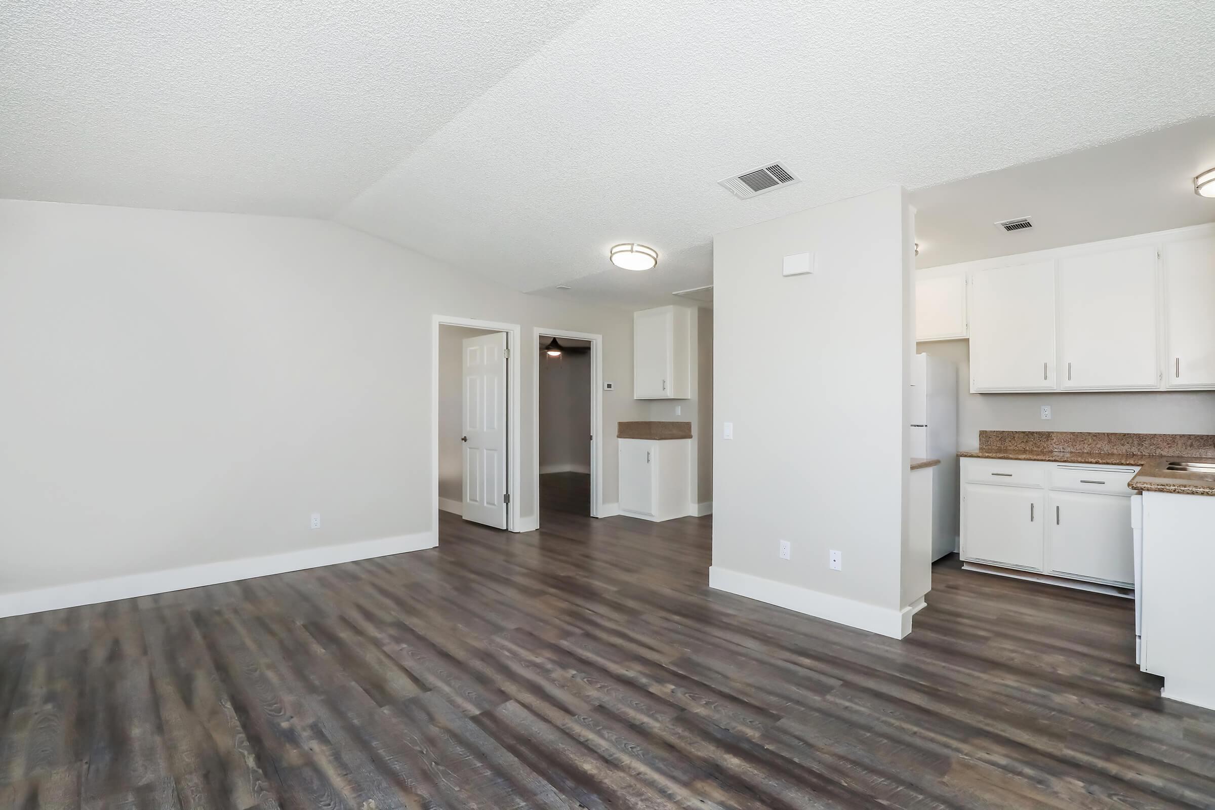 A spacious and modern living area featuring light-colored walls, hardwood-style flooring, and a kitchen bar. The kitchen is equipped with white cabinets and appliances, while doorways lead to other rooms. Ambient lighting fixtures add brightness to the room.