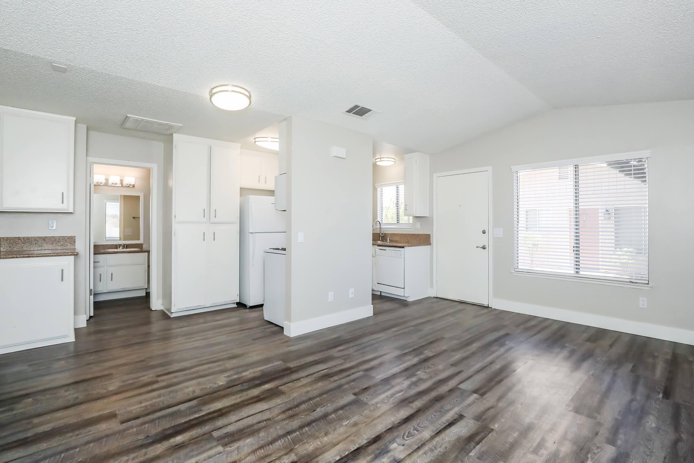 Spacious open-concept living area featuring modern kitchen appliances, including a refrigerator and white cabinetry. The room has large windows allowing natural light, and a door leads outside. The flooring is a warm, dark wood type, and the walls are painted in neutral tones, creating an inviting atmosphere.