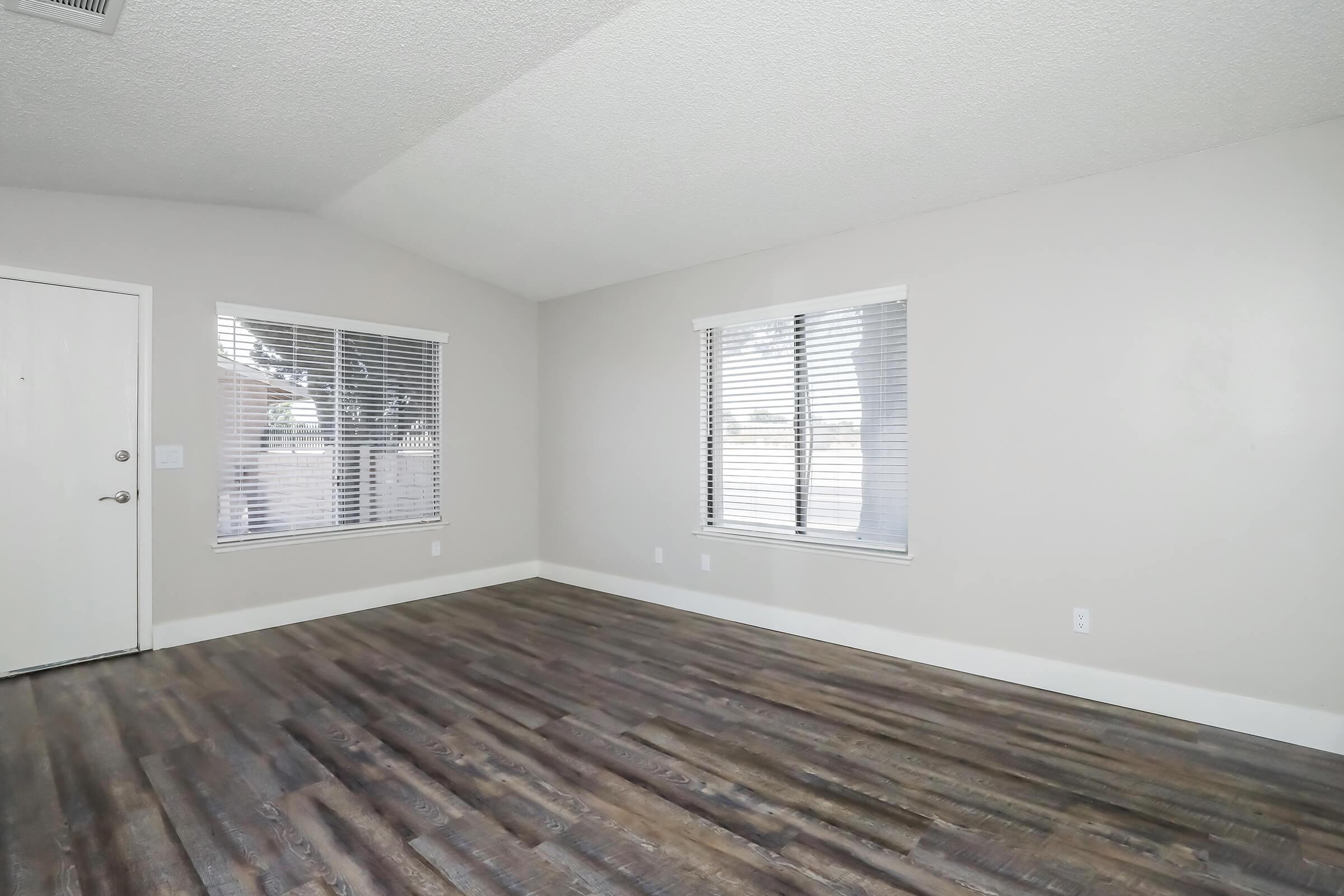 Spacious, empty living room featuring light gray walls, high ceilings, and large windows with white blinds. The floor consists of dark wood laminate, creating a warm contrast. A front door is visible on the left, while the room is bright and open, suitable for various furniture arrangements.