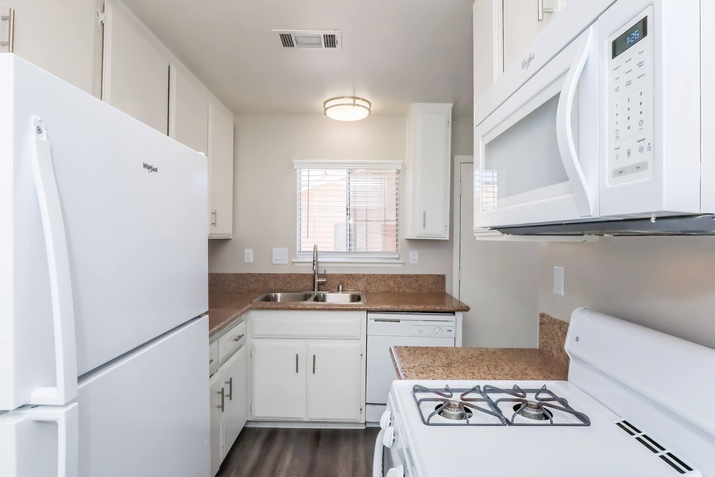 A modern kitchen featuring white appliances, including a refrigerator, microwave, and stove. The countertops are a light brown with a sink underneath a window. Light cabinets provide storage, and the flooring is a dark wood laminate, creating a clean and functional space.