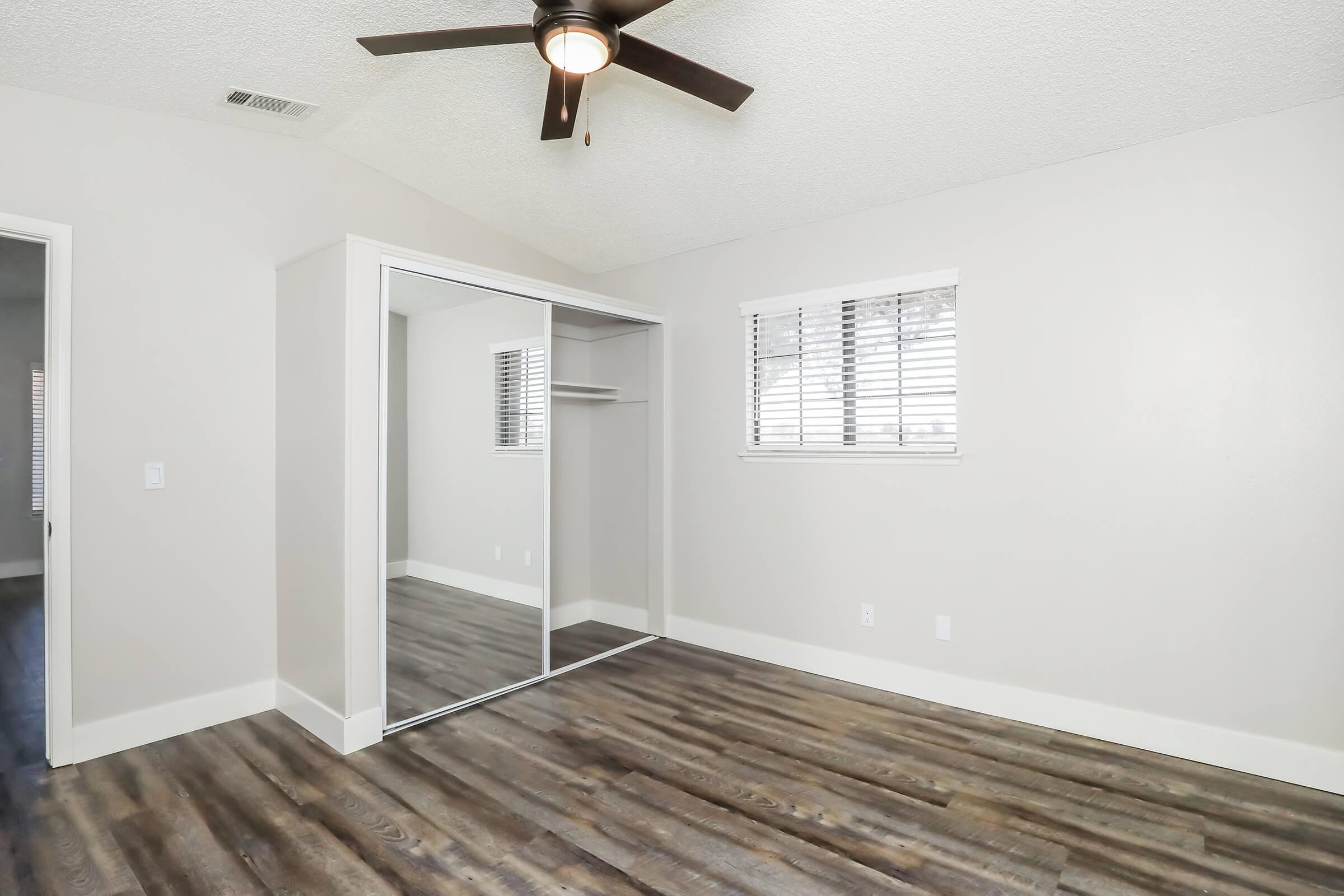 A well-lit room featuring a ceiling fan, large mirrored wardrobe, and beige walls. The room has wooden flooring and a window with white blinds. There is an entrance to another space on the left, enhancing the overall spaciousness and modern design.