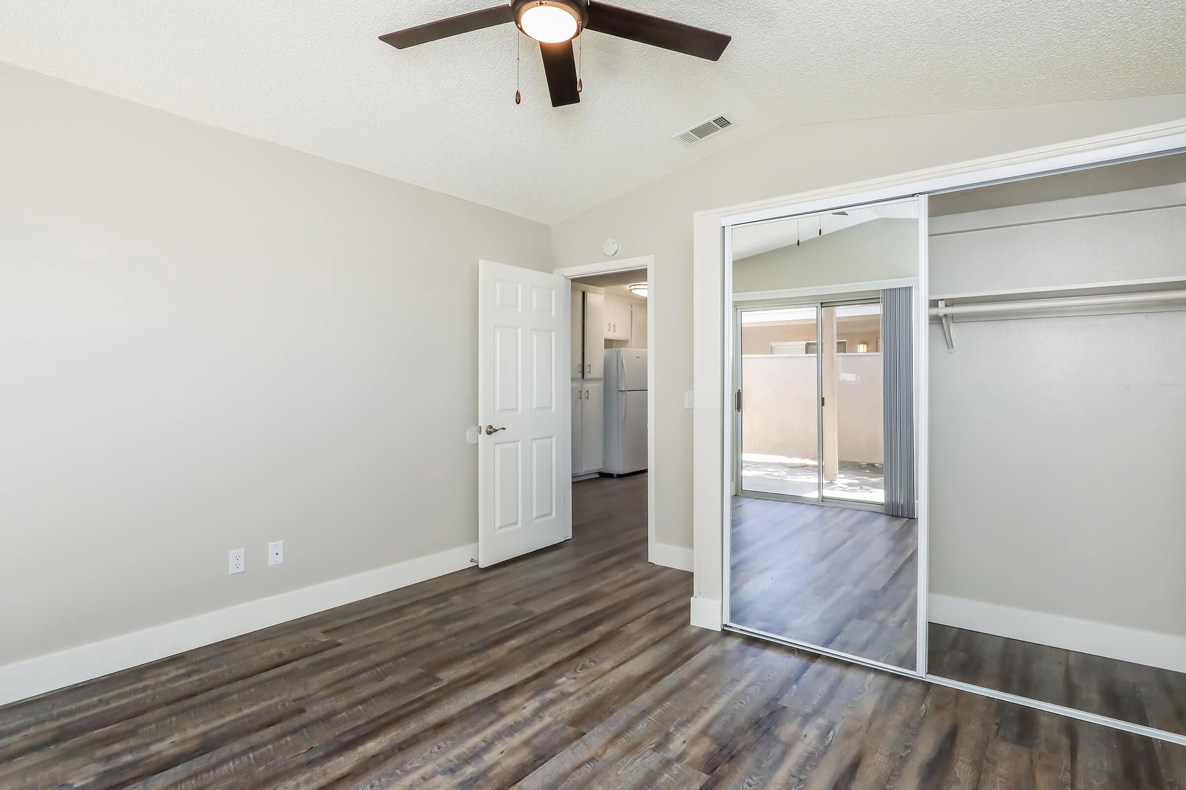A bright room with a ceiling fan, light gray walls, and wooden flooring. There is a doorway leading to another area and a large mirrored closet. The space is well-lit, featuring a window that opens to a view outside. The room has a clean and modern design, suitable for various uses.