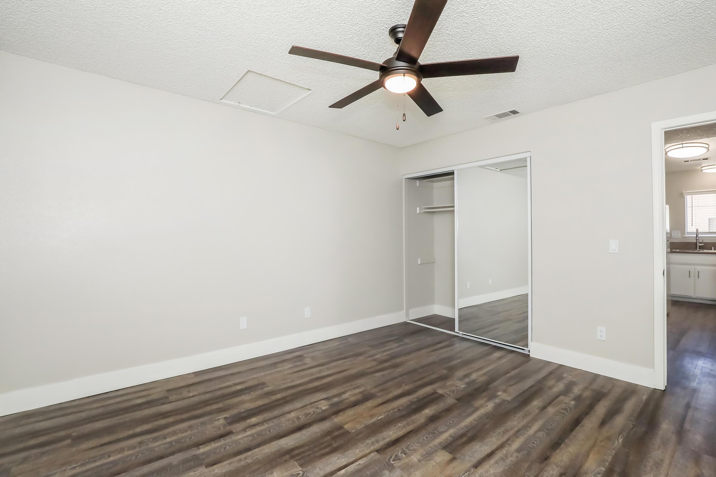 A spacious room with light-colored walls and a ceiling fan. The floor features dark wood-like laminate. There's a mirrored closet with sliding doors on one side and a doorway leading to another room or area in the background. Natural light comes in from a nearby window.